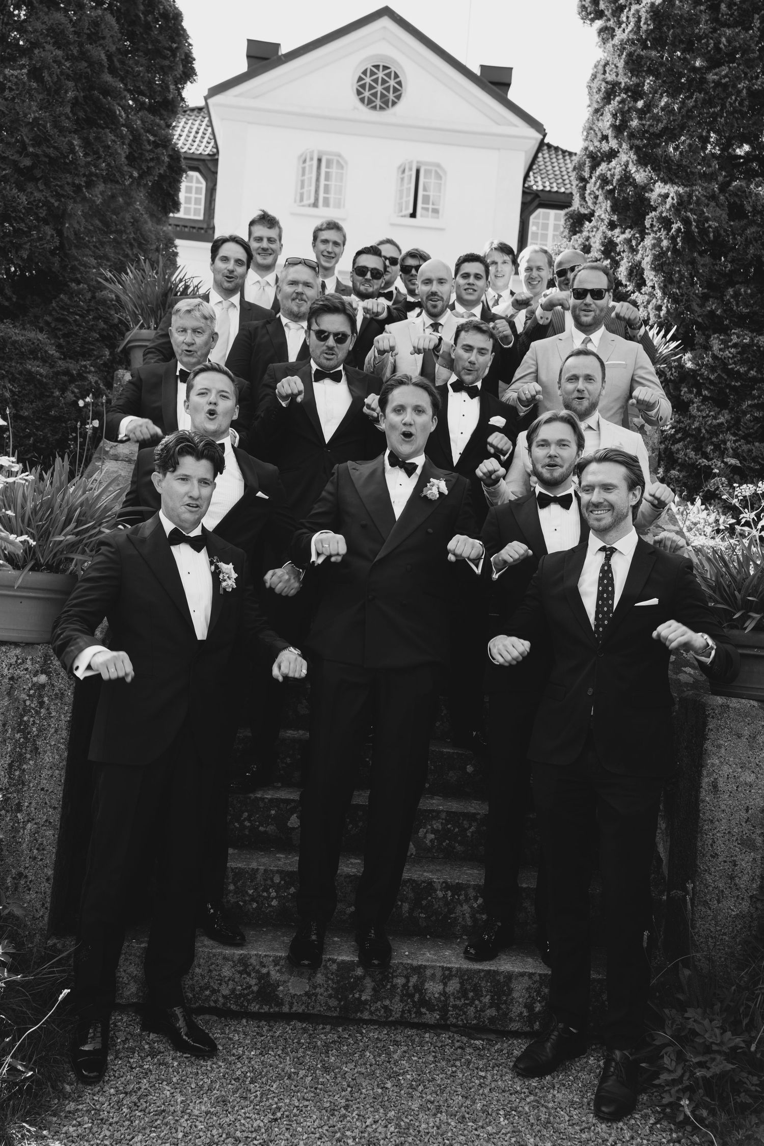 Black and white group portrait of groom with groomsmen in tuxedos posing playfully on stone steps outside a manor house.