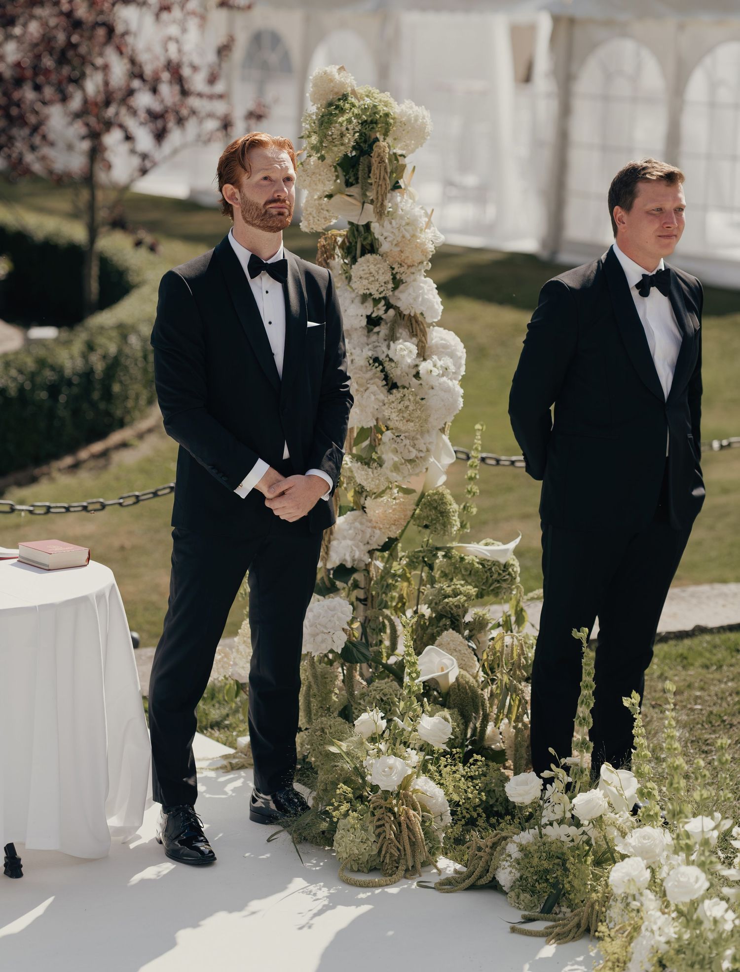 Two groomsmen in black tuxedos and bow ties stand beside a tall white floral arch at an outdoor garden wedding ceremony.