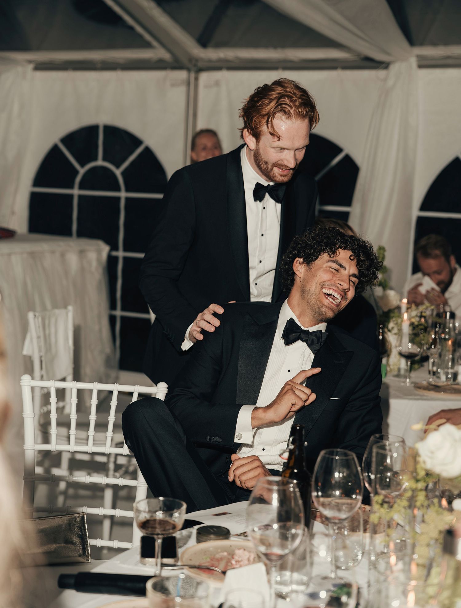 Groom in a black tuxedo laughs joyfully at a wedding reception table while a smiling groomsman rests a hand on his shoulder.
