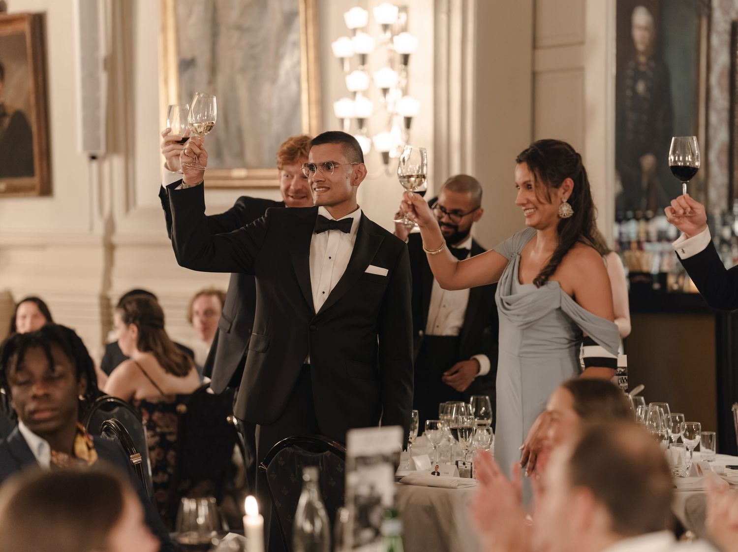 Guests in formal attire raise champagne glasses in a toast at an elegant banquet hall with chandeliers and set dining tables.