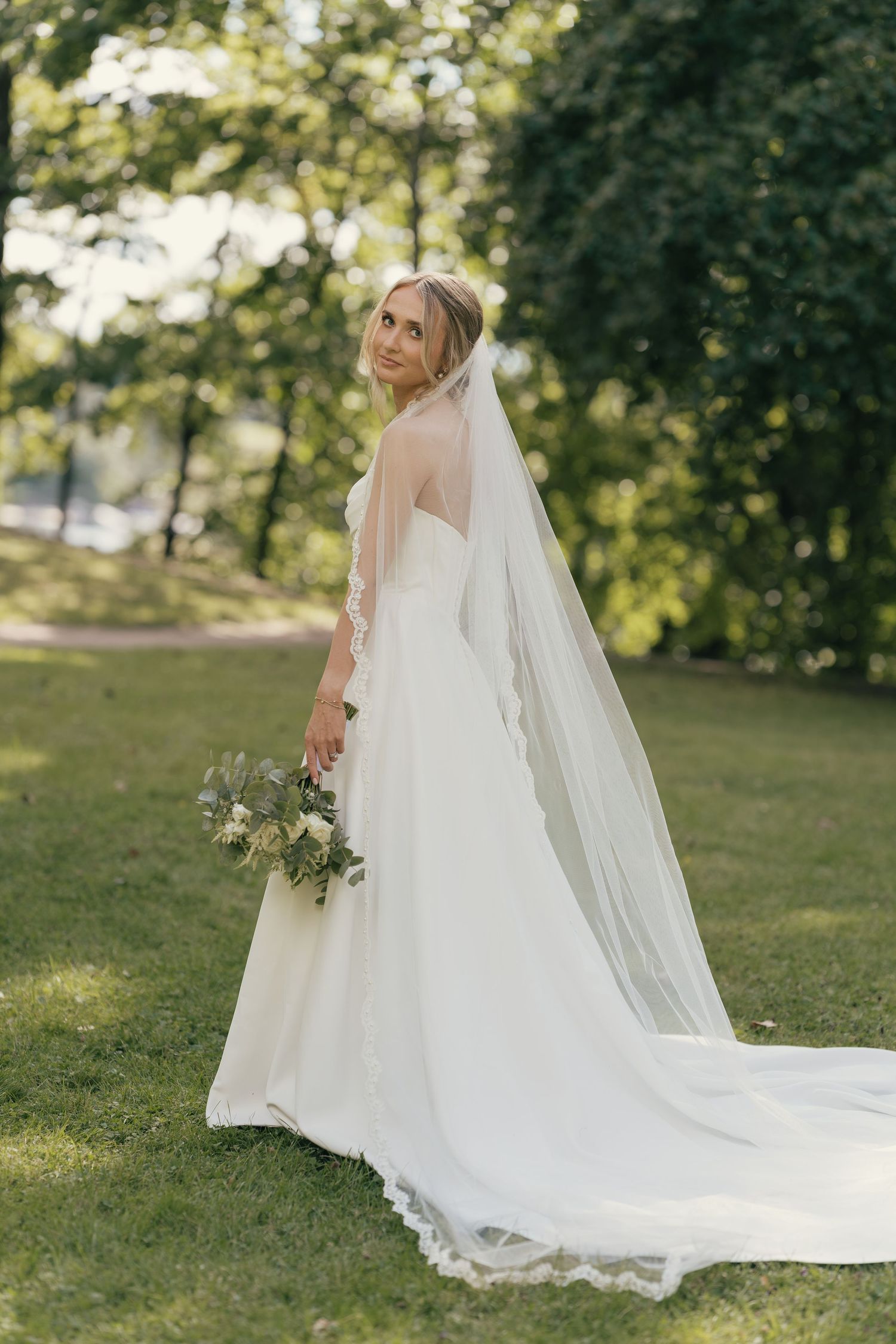 Bride in white gown and long veil holds bouquet, standing on green grass in a sunlit park.