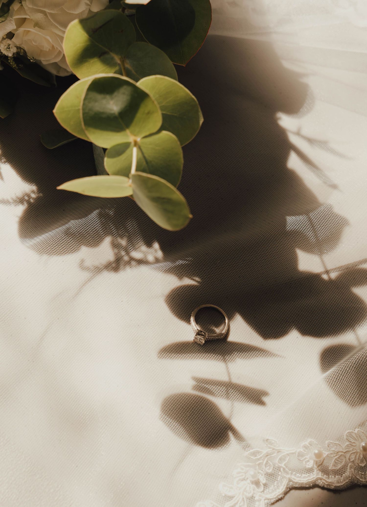 Wedding ring resting on bridal veil beside green eucalyptus leaves in warm sunlight.