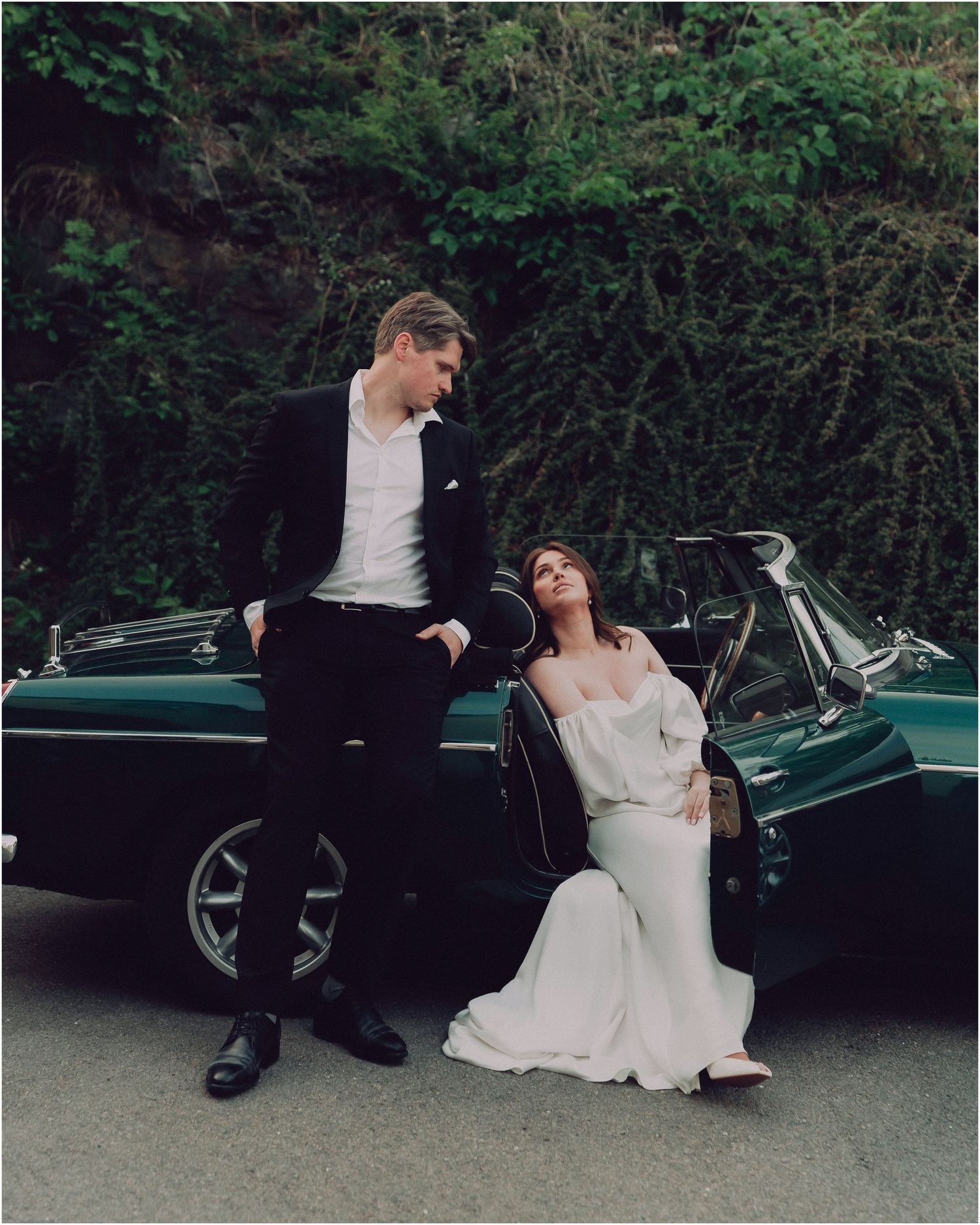 A couple in wedding attire sits and laughs together next to a vintage green car on a tree-lined road.