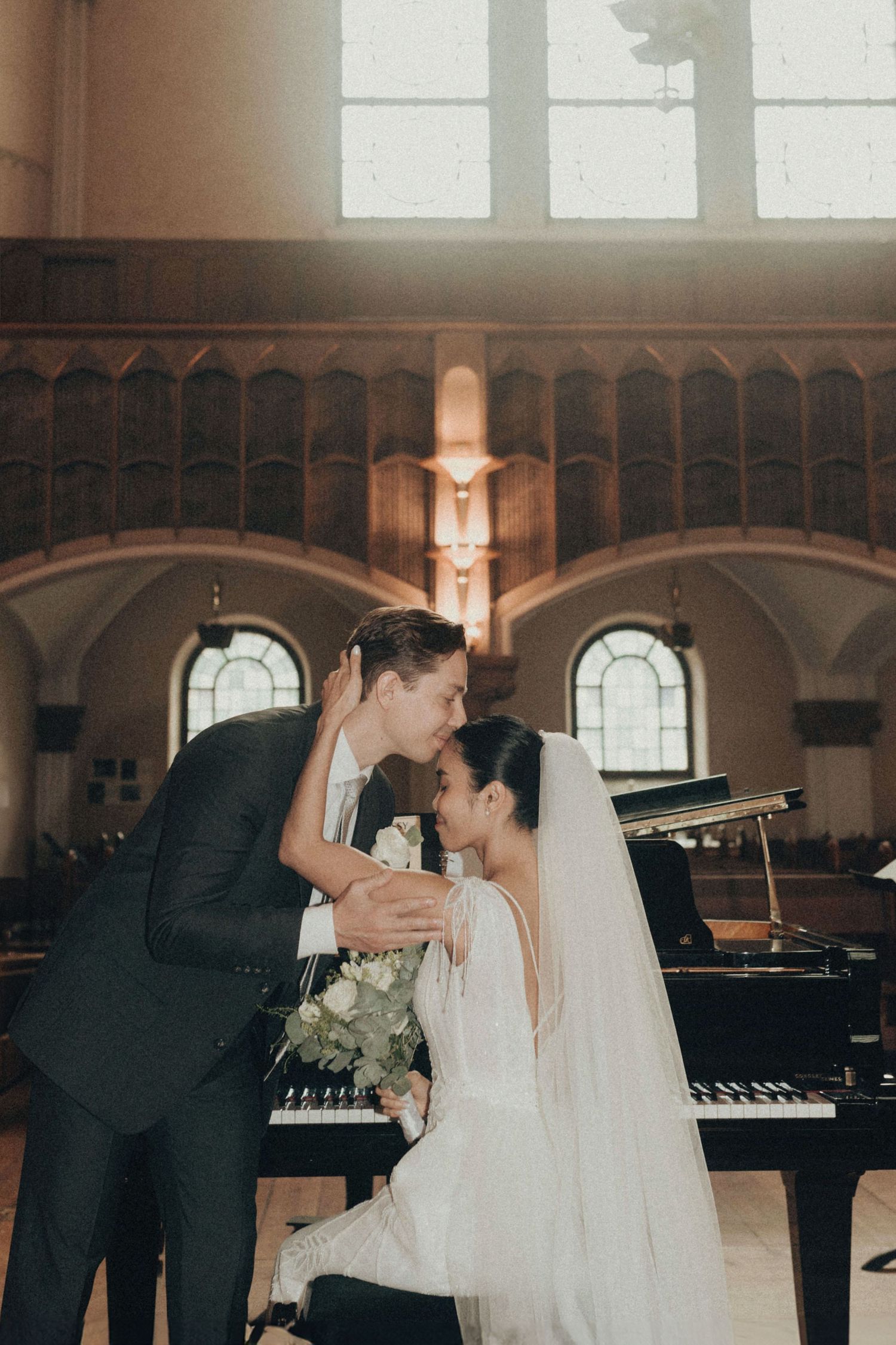 Romantic wedding couple shares tender moments by a grand piano in an elegant church venue with arched windows.