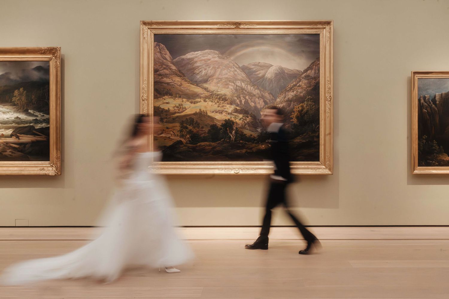 Blurred bride and groom walking past large landscape paintings in an art museum during their wedding.