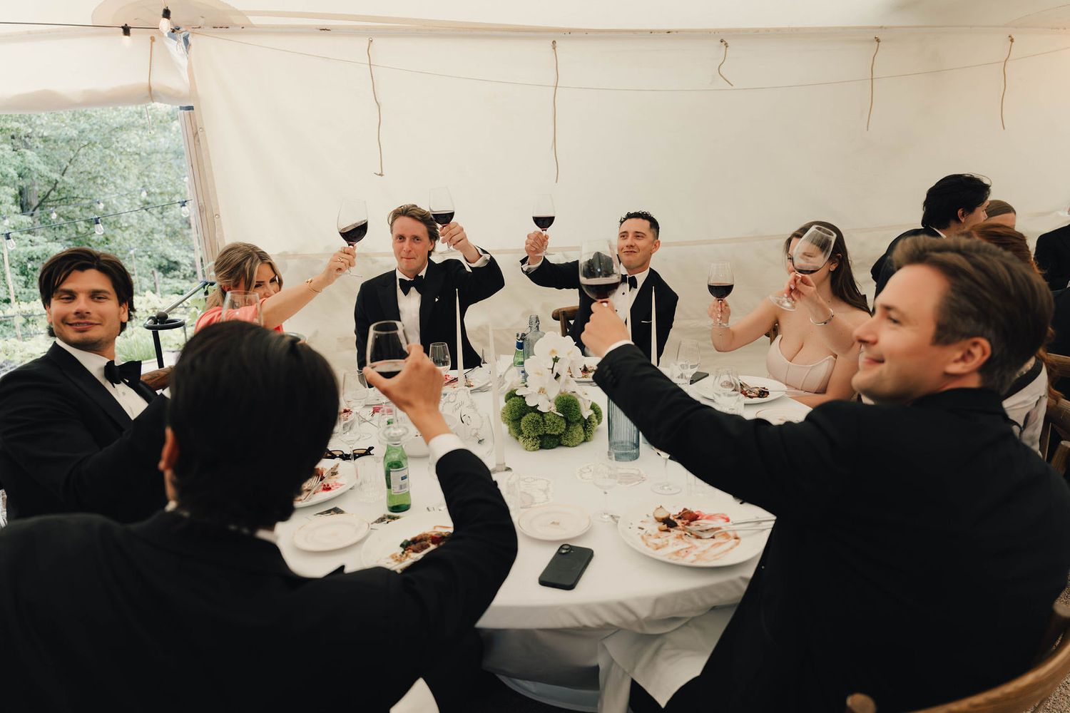 Wedding guests in formal attire raising glasses of red wine in a celebratory toast at an elegant reception.