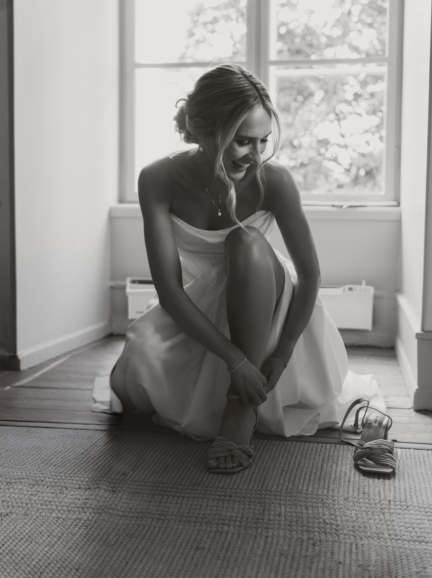 Bride in white strapless dress sitting on floor fastening strappy sandals near window in black and white photo.