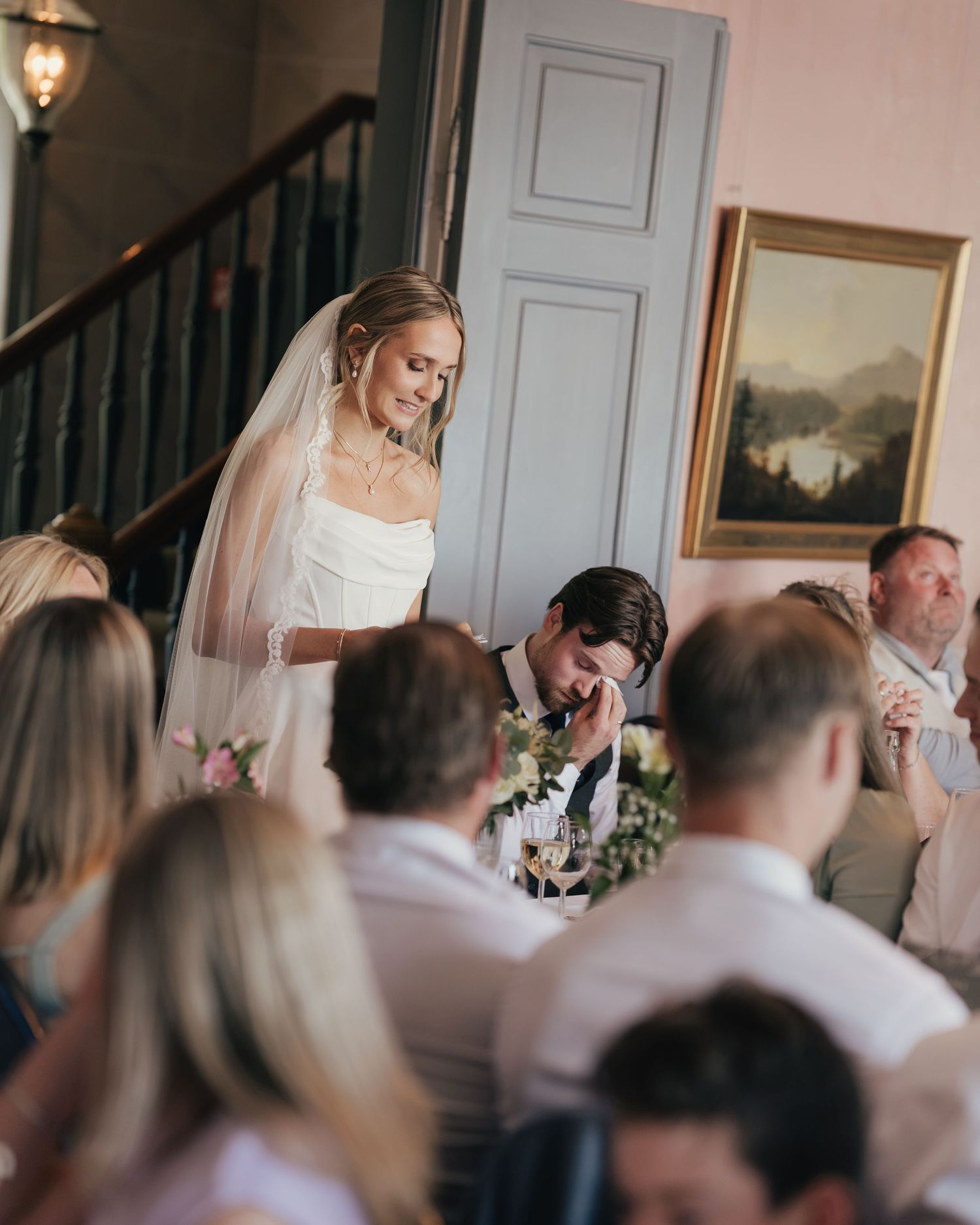 Bride in white gown and veil leans over emotional groom during wedding reception speeches.