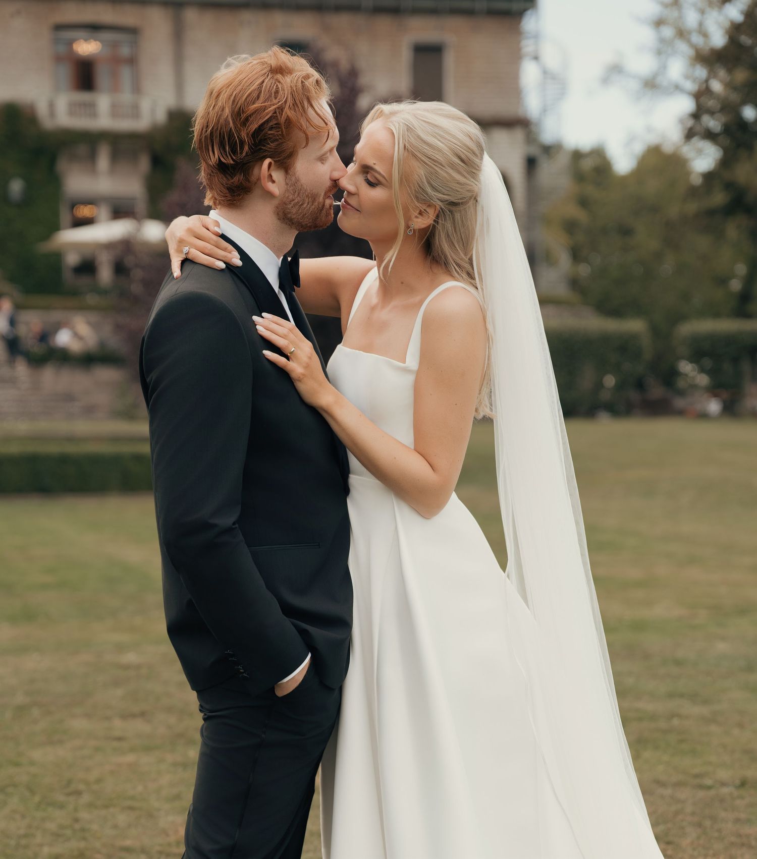 Groom in a dark suit kisses a bride in a white gown and long veil on a lush green lawn with a historic building behind.