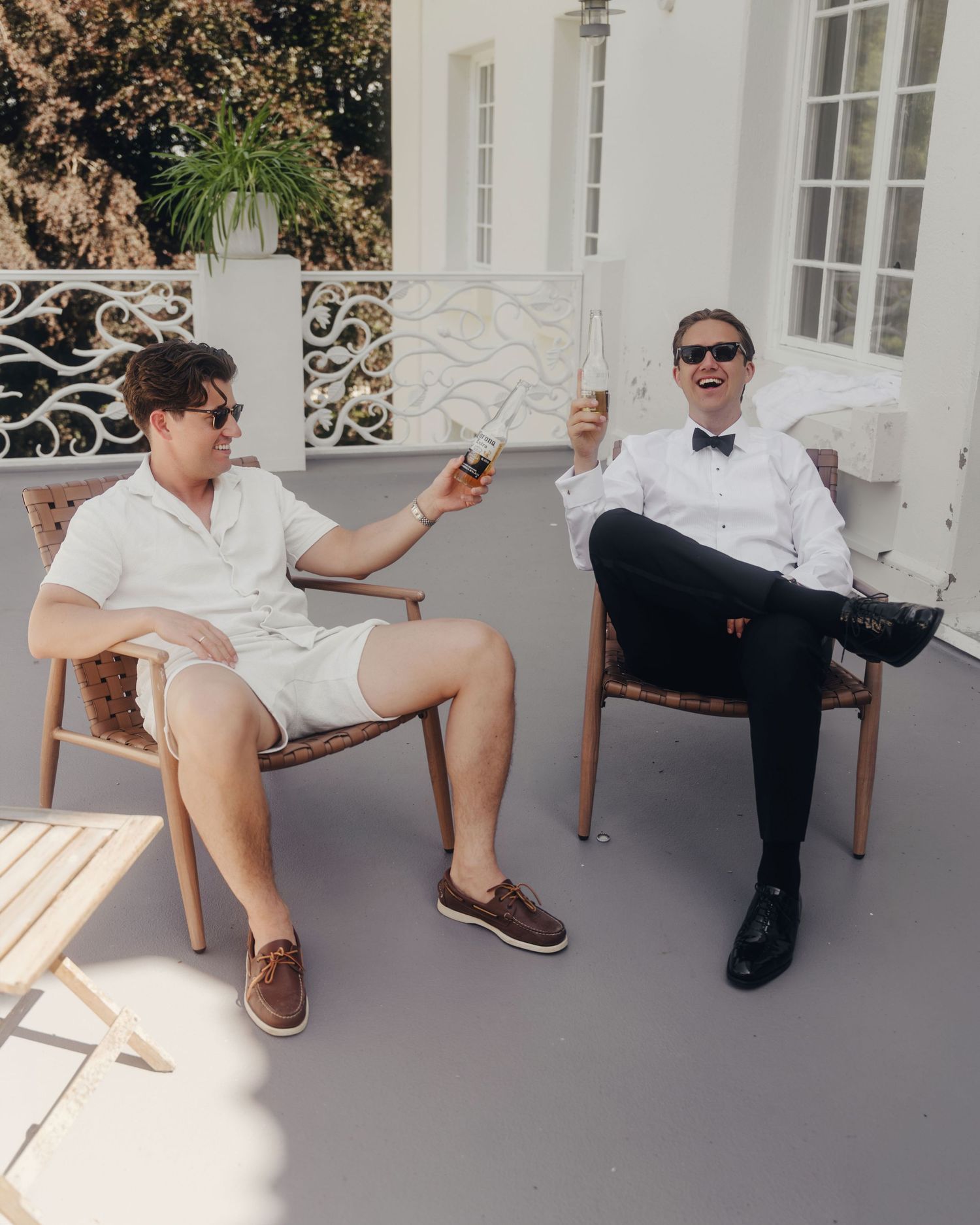 Two men in sunglasses laugh and hold drinks while relaxing in wooden chairs on a sunny terrace, one in casual shorts and one in a bow tie.