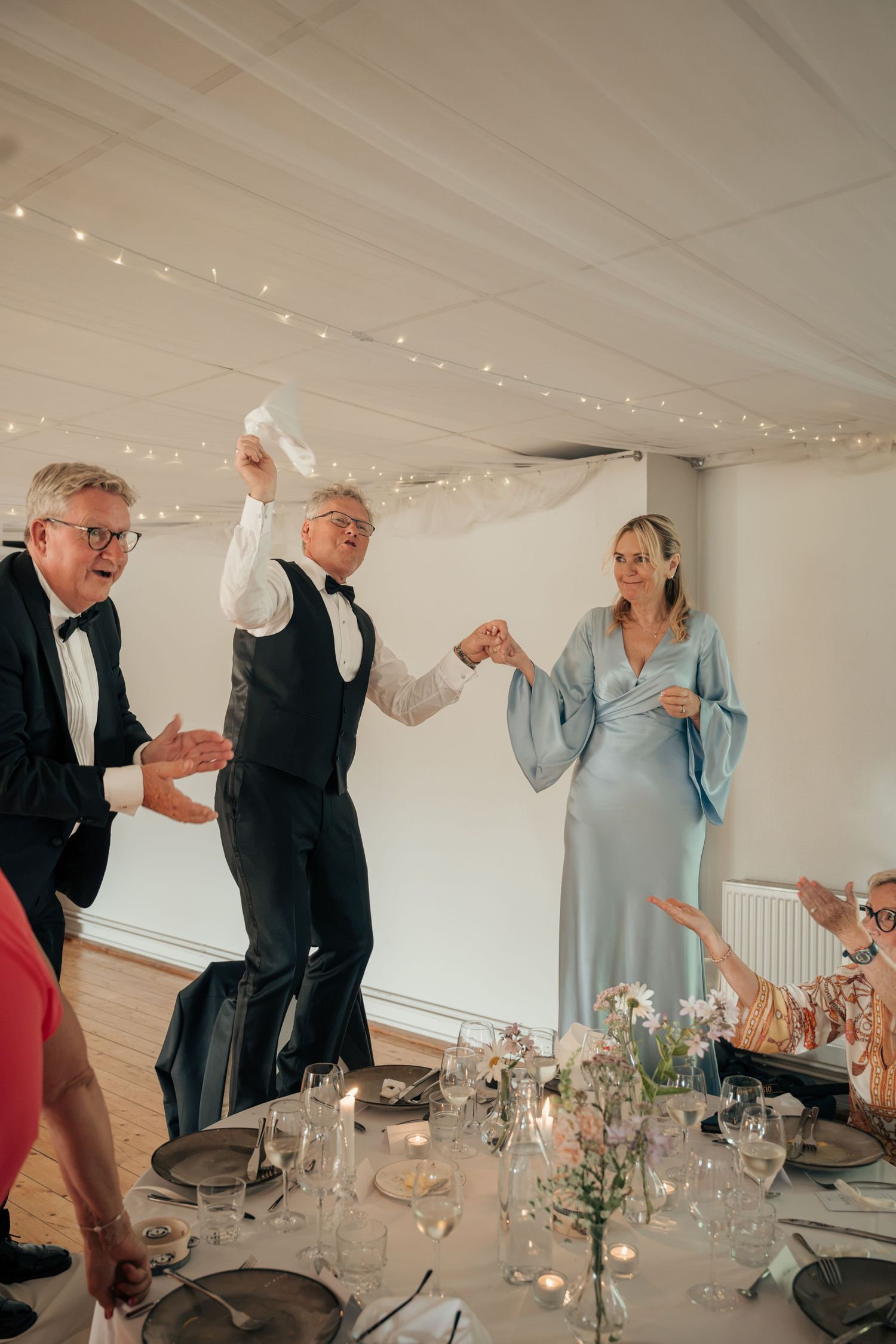 Wedding guests dance and wave napkins near a reception table as a woman in a flowing blue dress joins hands with a man.