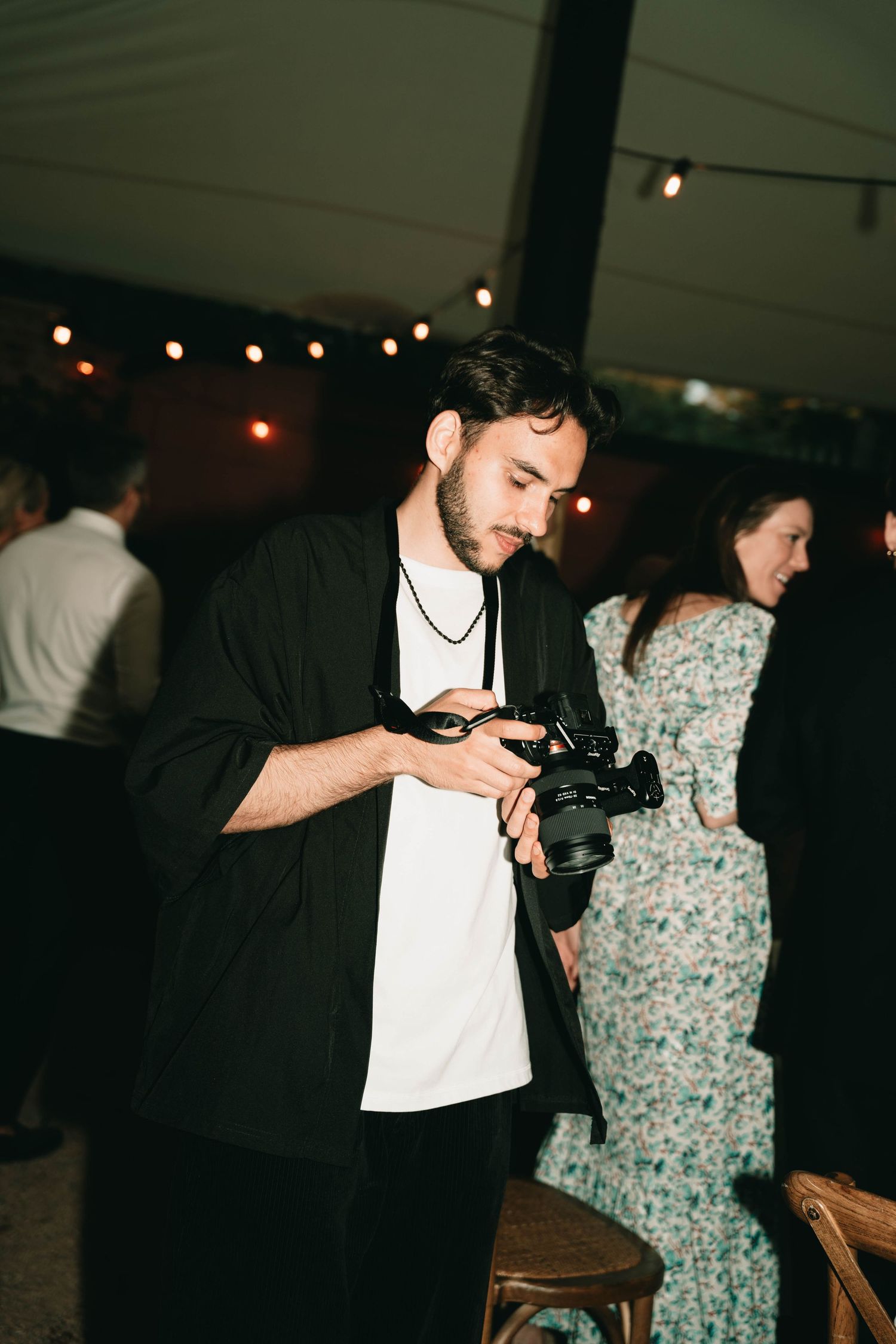 A person in a black jacket and white shirt takes a photo with a vintage camera under string lights at an evening event.