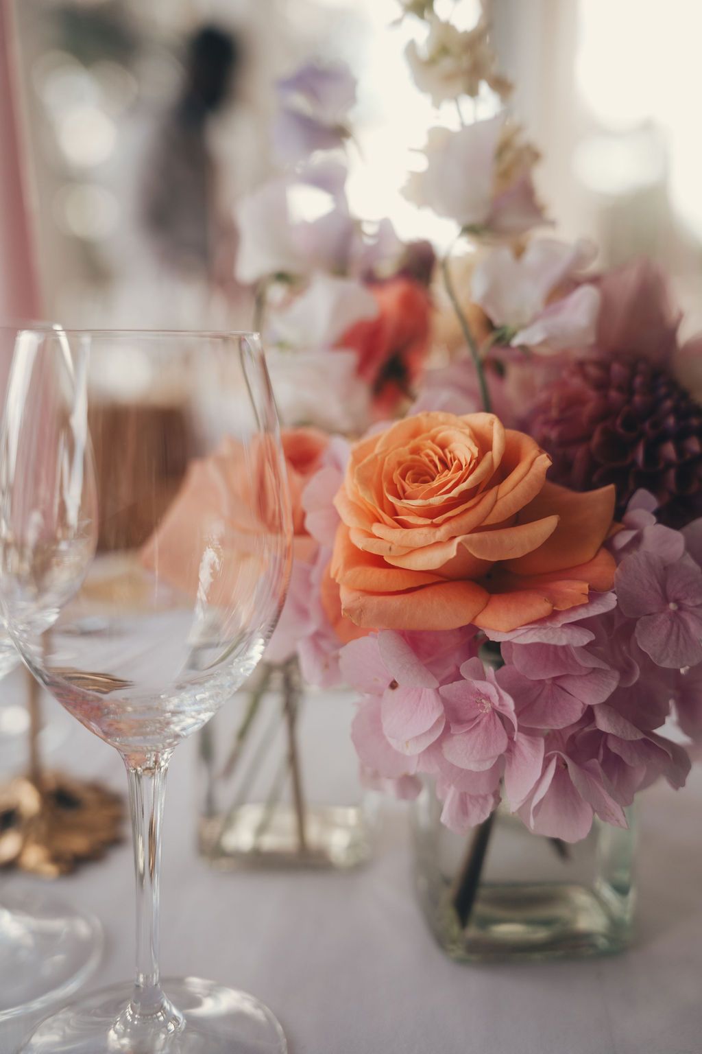 Elegant wedding table setting with peach roses, pink hydrangeas, and crystal wine glasses in soft natural light.