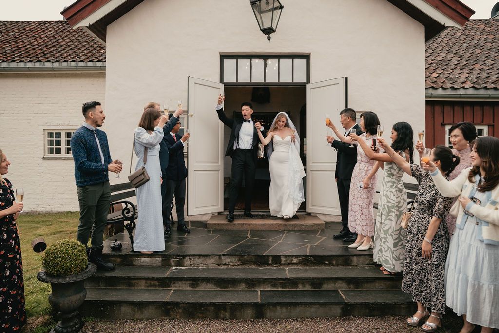 Wedding party stands on stone steps outside rustic building with tiled roof in natural light.