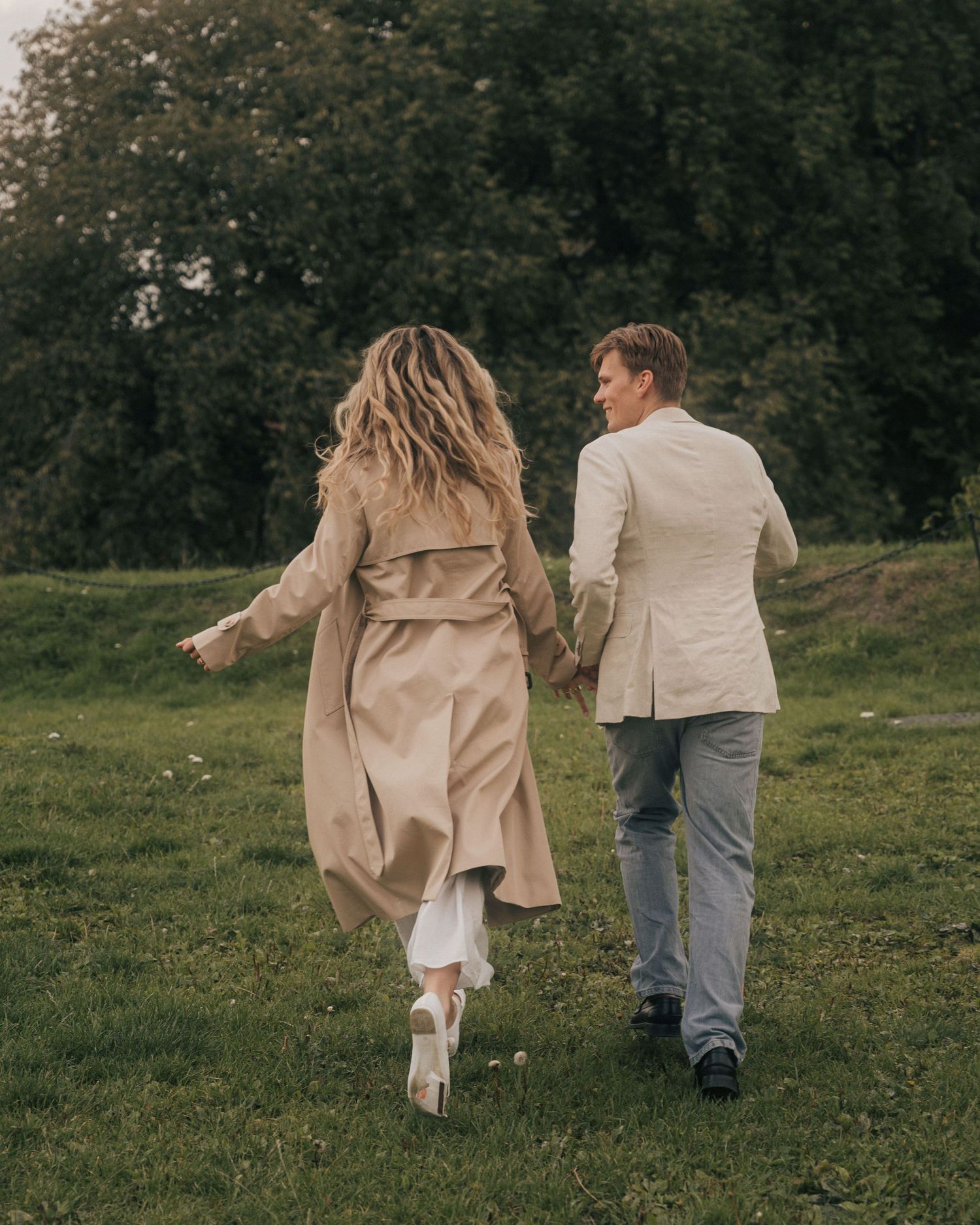 A couple in wedding attire walks hand in hand through a lush green meadow on an overcast day.