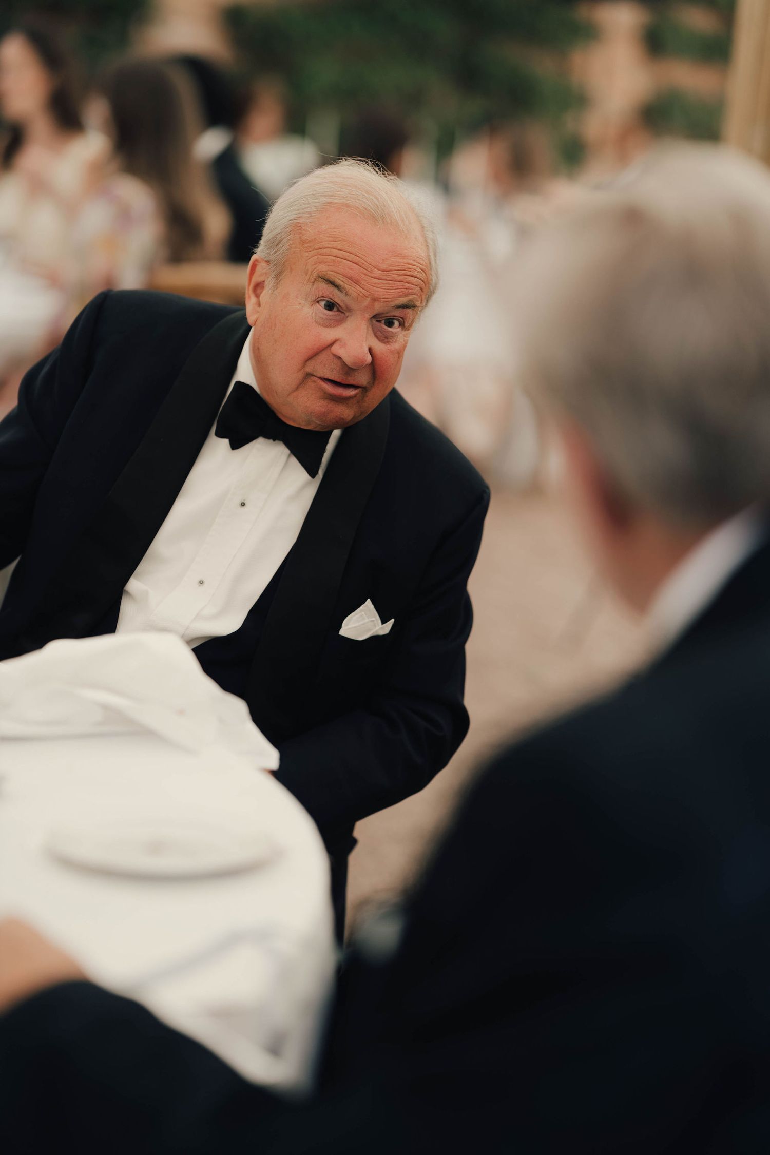 Elderly man in tuxedo with bow tie engaged in conversation at formal event.