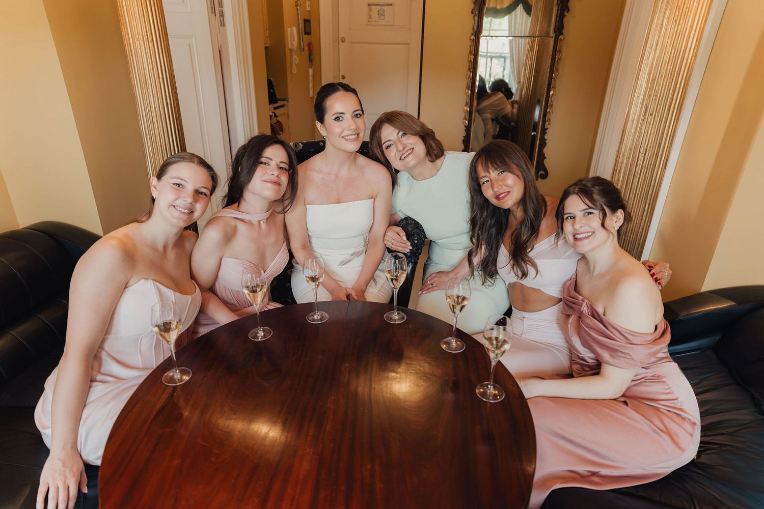 Bride with bridesmaids in pink dresses toasting with champagne glasses around a wooden table at a wedding.