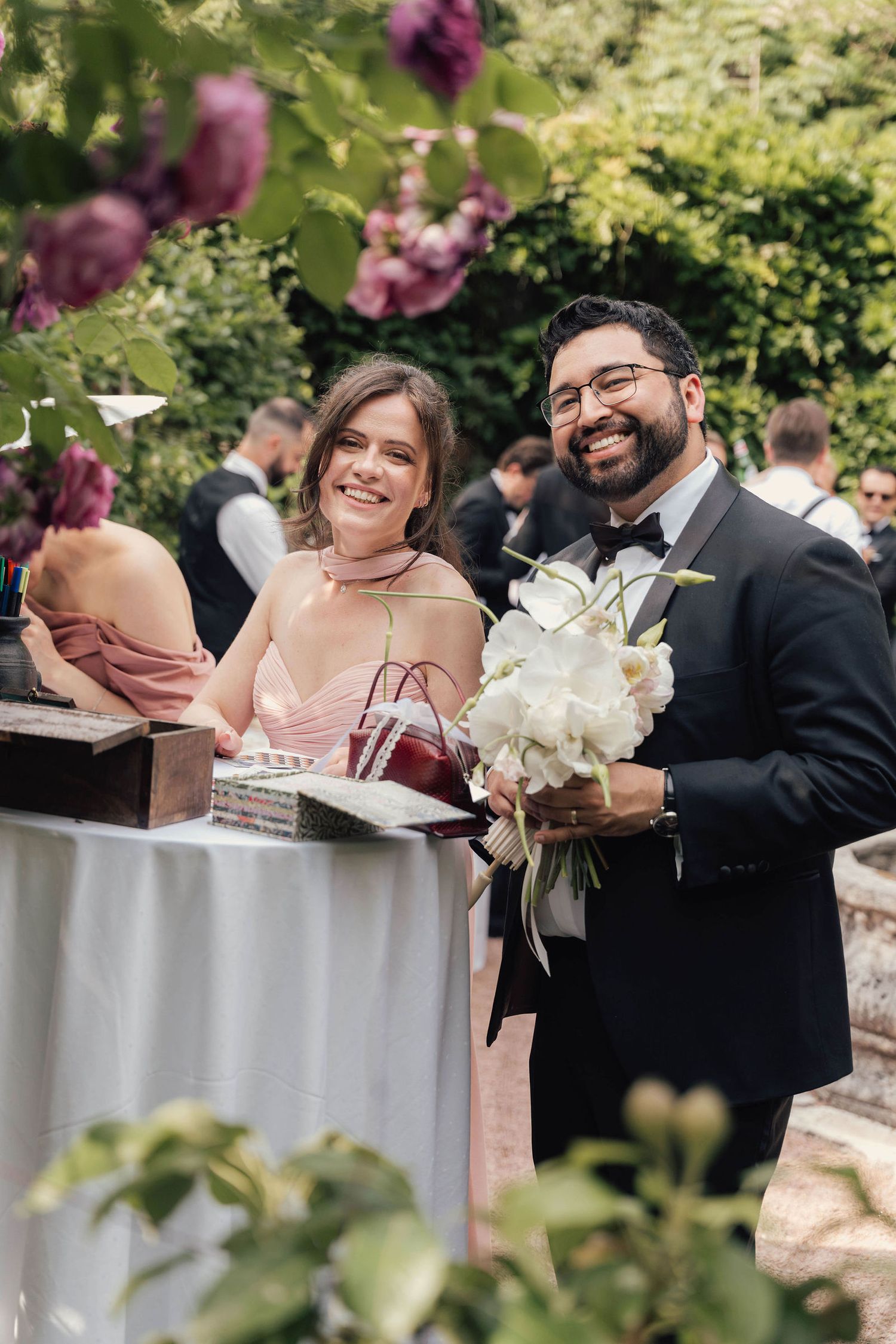 Smiling couple at outdoor wedding, man in tuxedo holding white bouquet, woman in pink dress, garden setting.