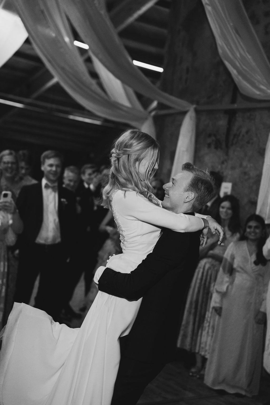 Black and white photo of couples dancing at an elegant reception with draped fabric decor and guests watching.