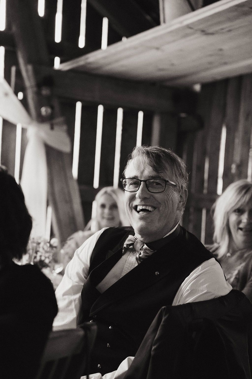 Black and white candid photo of a guest laughing during a reception in a rustic barn setting.