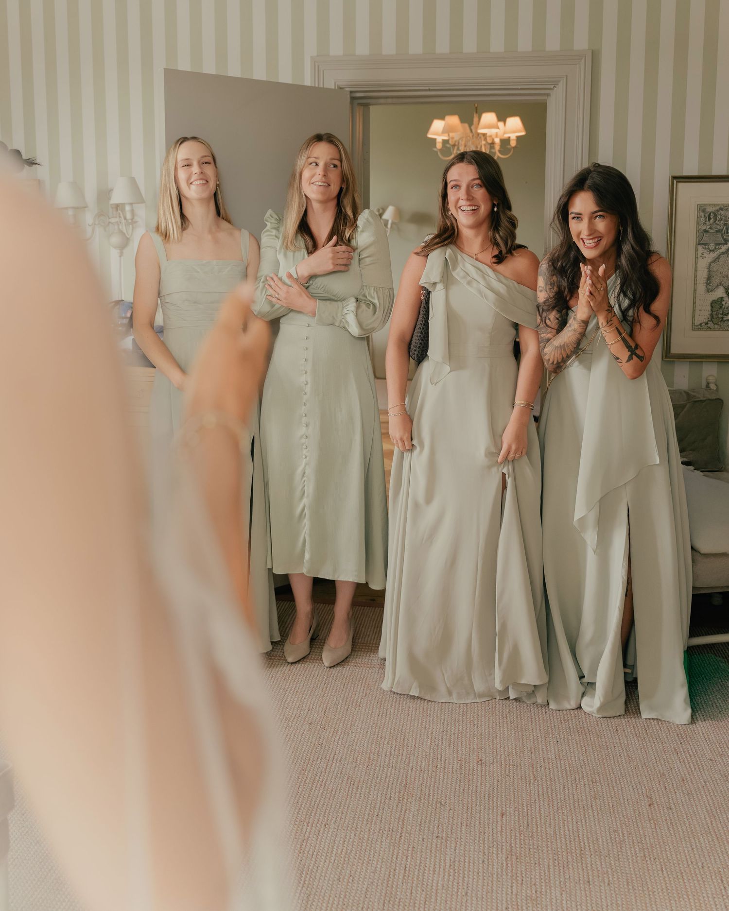 Four bridesmaids in sage green satin gowns react with joy upon seeing the bride during wedding preparations.
