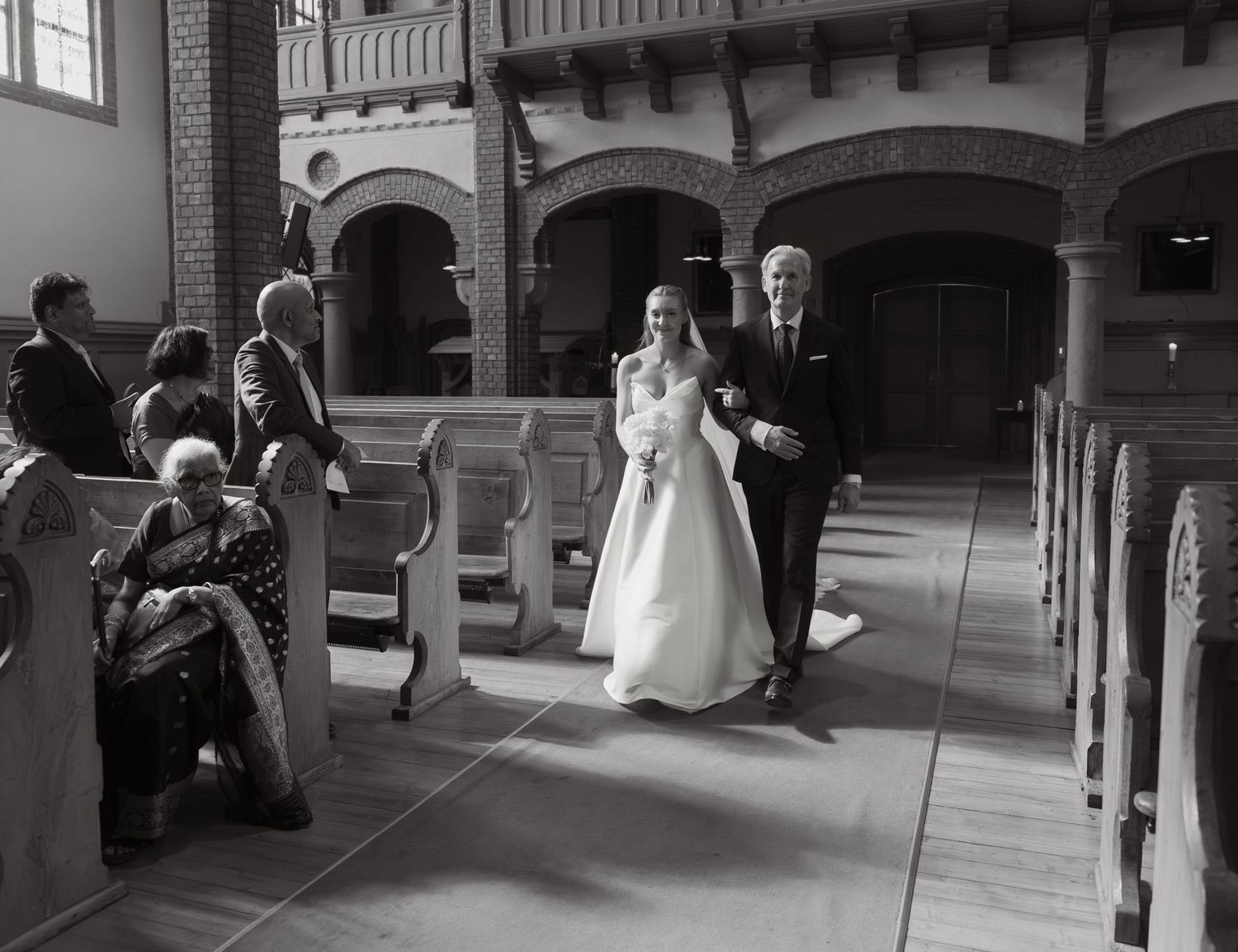 Black and white photo of a bride in a lace gown walking down the church aisle on a man's arm as seated wedding guests look on.