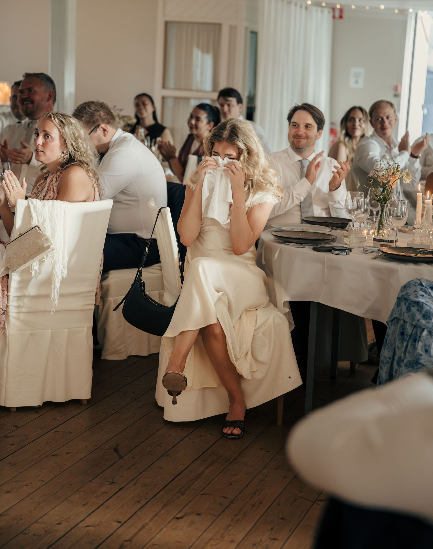 Emotional bride in a white dress dabs her eyes with a napkin while seated among applauding wedding reception guests.