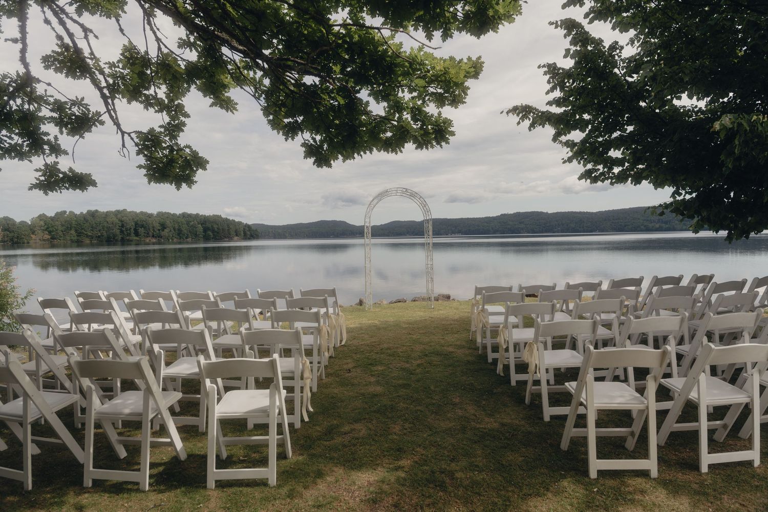 White folding chairs set in rows on a grassy lawn facing a wedding arch beside a calm lake.