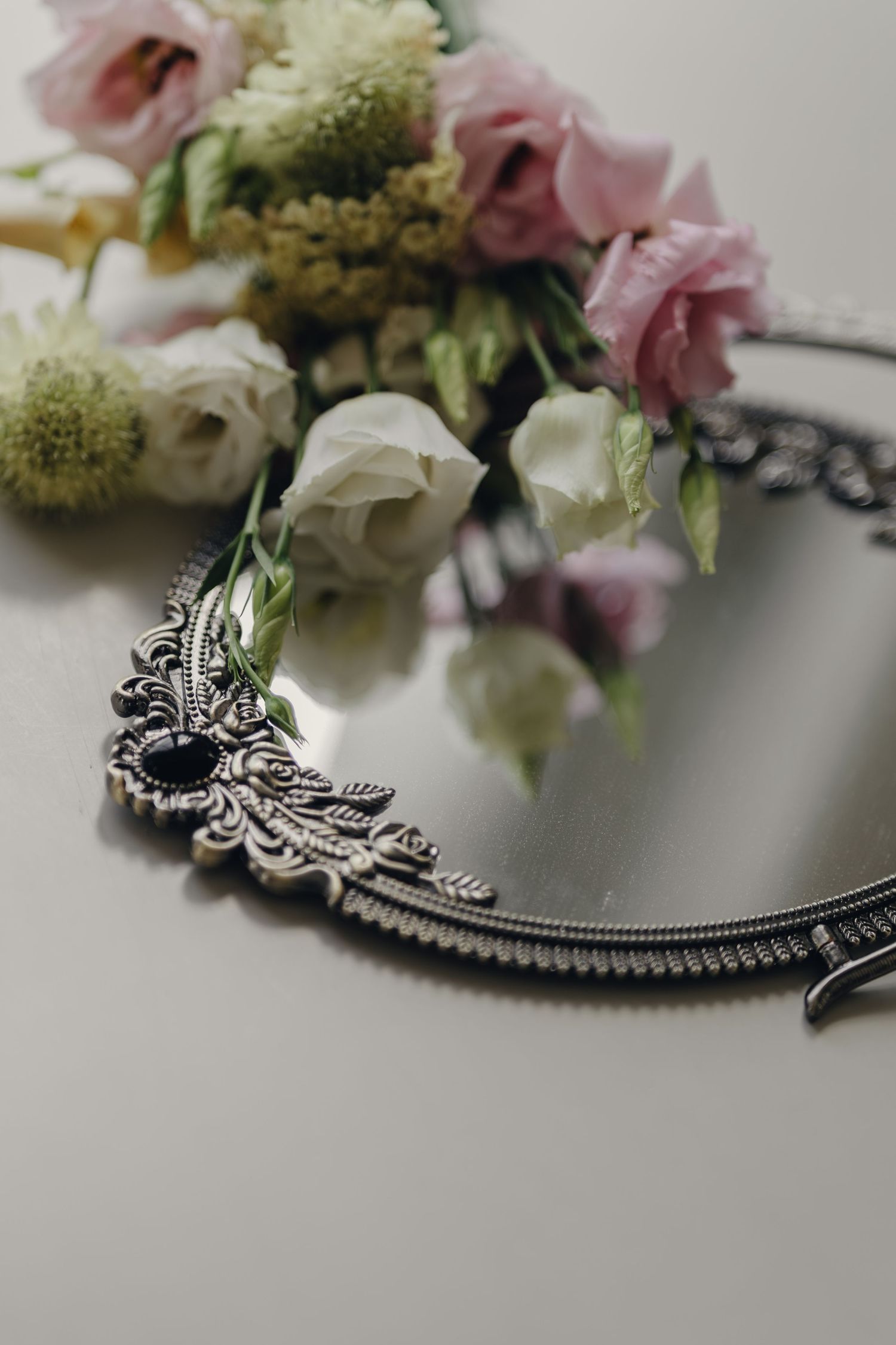 Pink and white lisianthus flowers with green sprigs resting on an ornate vintage silver mirror with intricate filigree detailing.