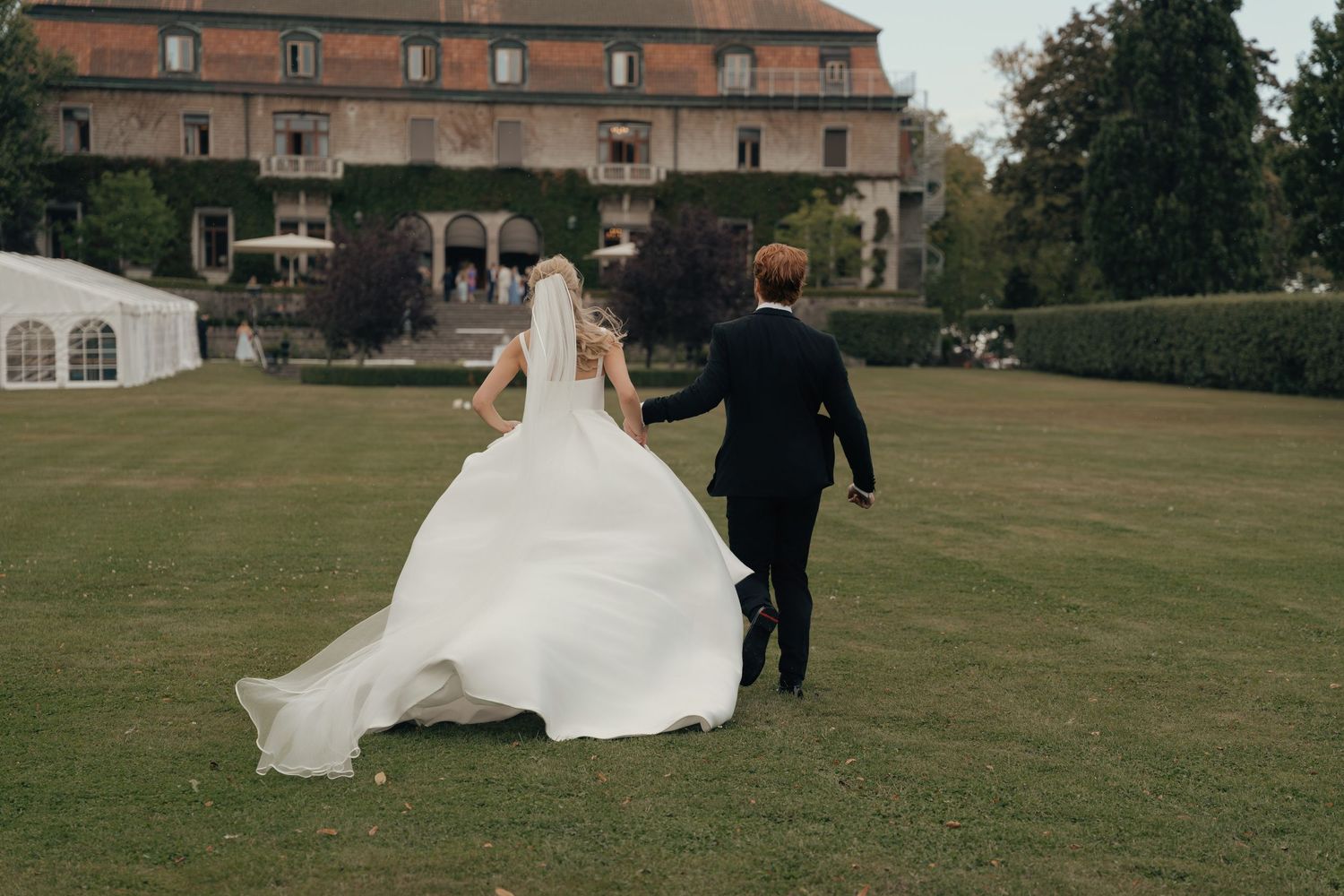Bride in a flowing white gown and groom in a dark suit walk hand in hand across a green lawn toward a historic manor.