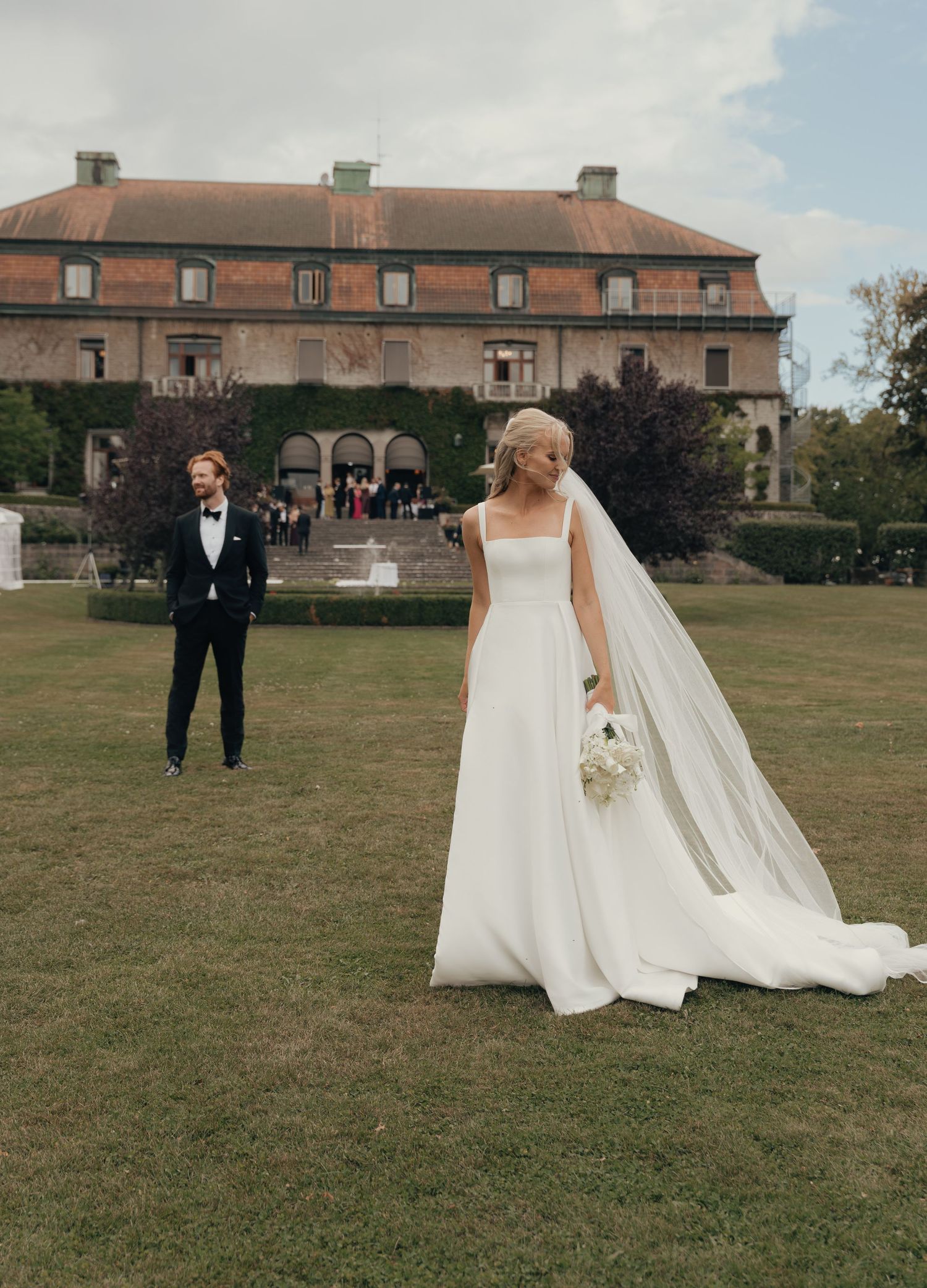 Bride in a white gown and long veil holds a bouquet on a lawn as the groom watches before a historic manor.