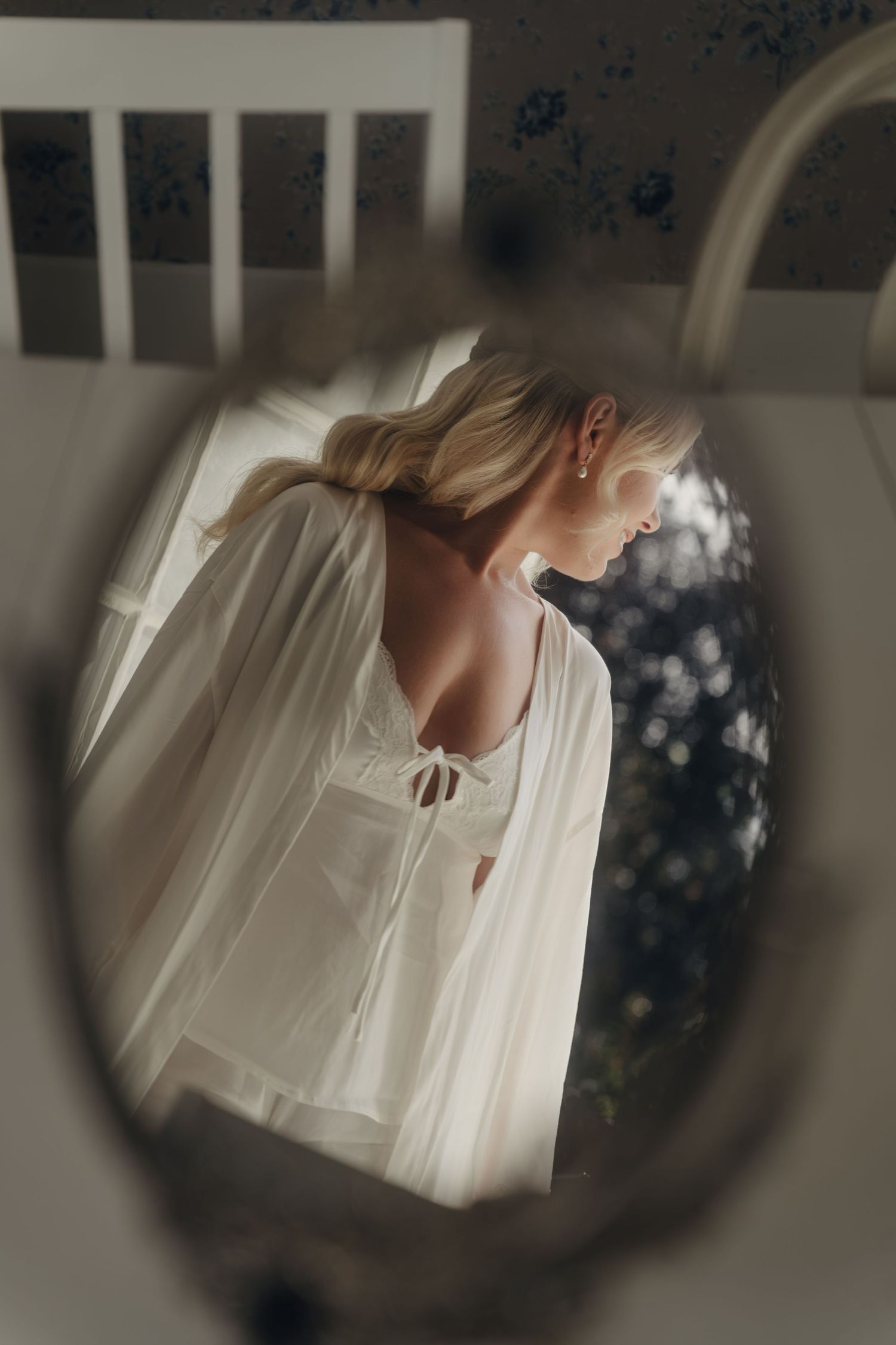 Reflection in a vintage oval mirror of a blonde woman in a white lace-up top and sheer robe, softly lit in a romantic bedroom.