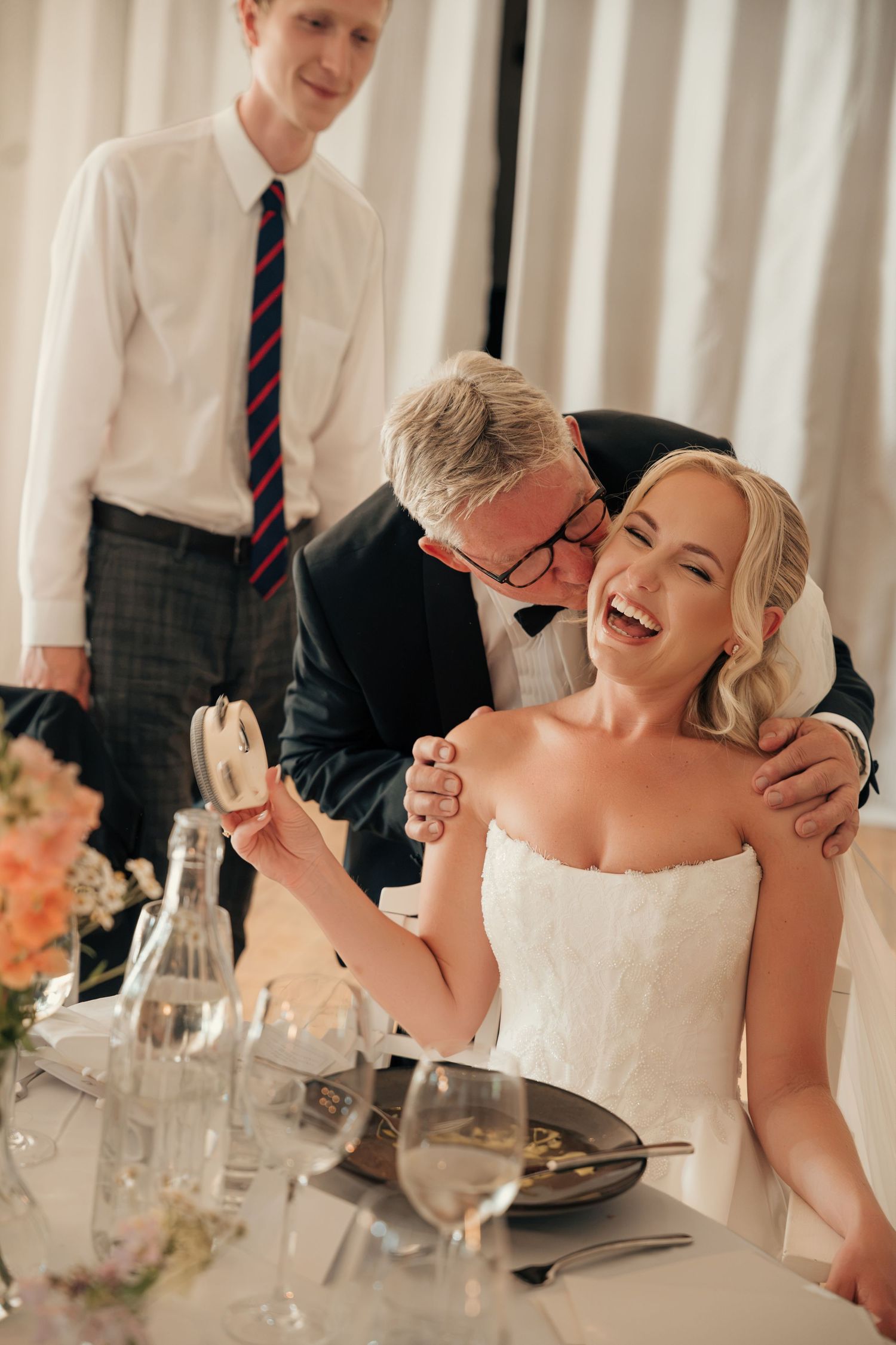 A laughing bride in a strapless white gown is kissed on the cheek by a man in glasses at a reception table as a guest looks on.