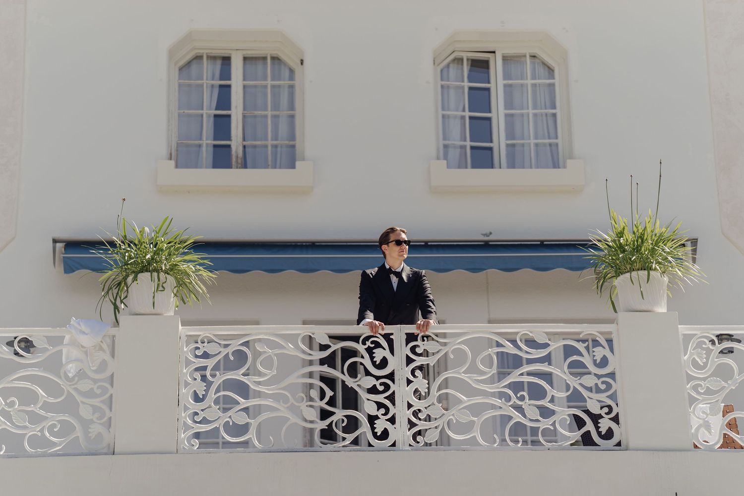 Person in dark suit and sunglasses stands on an ornate white balcony with potted plants beneath a blue awning and shuttered windows.