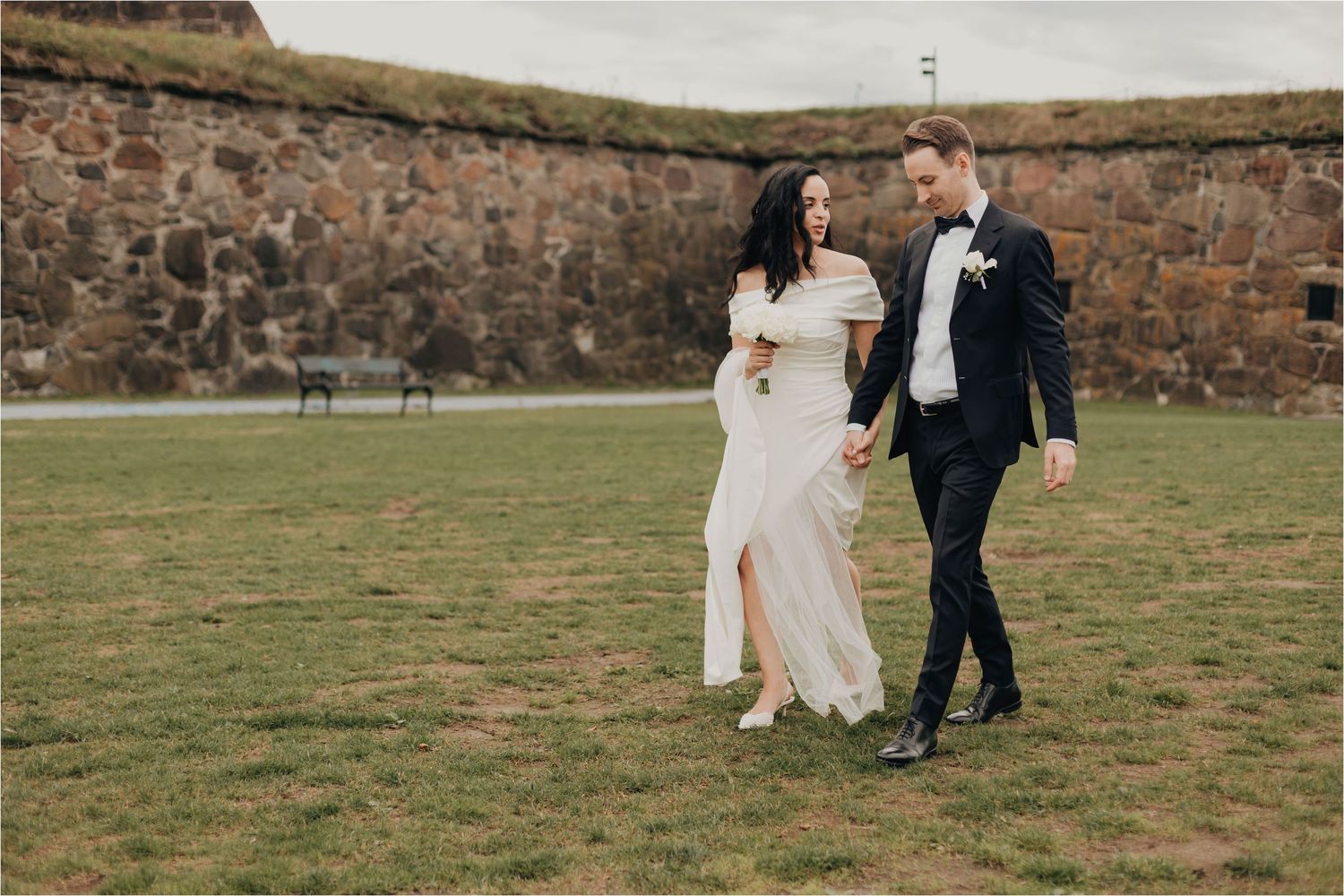 A couple walks together across a grassy field with stone walls in the background on their wedding day.