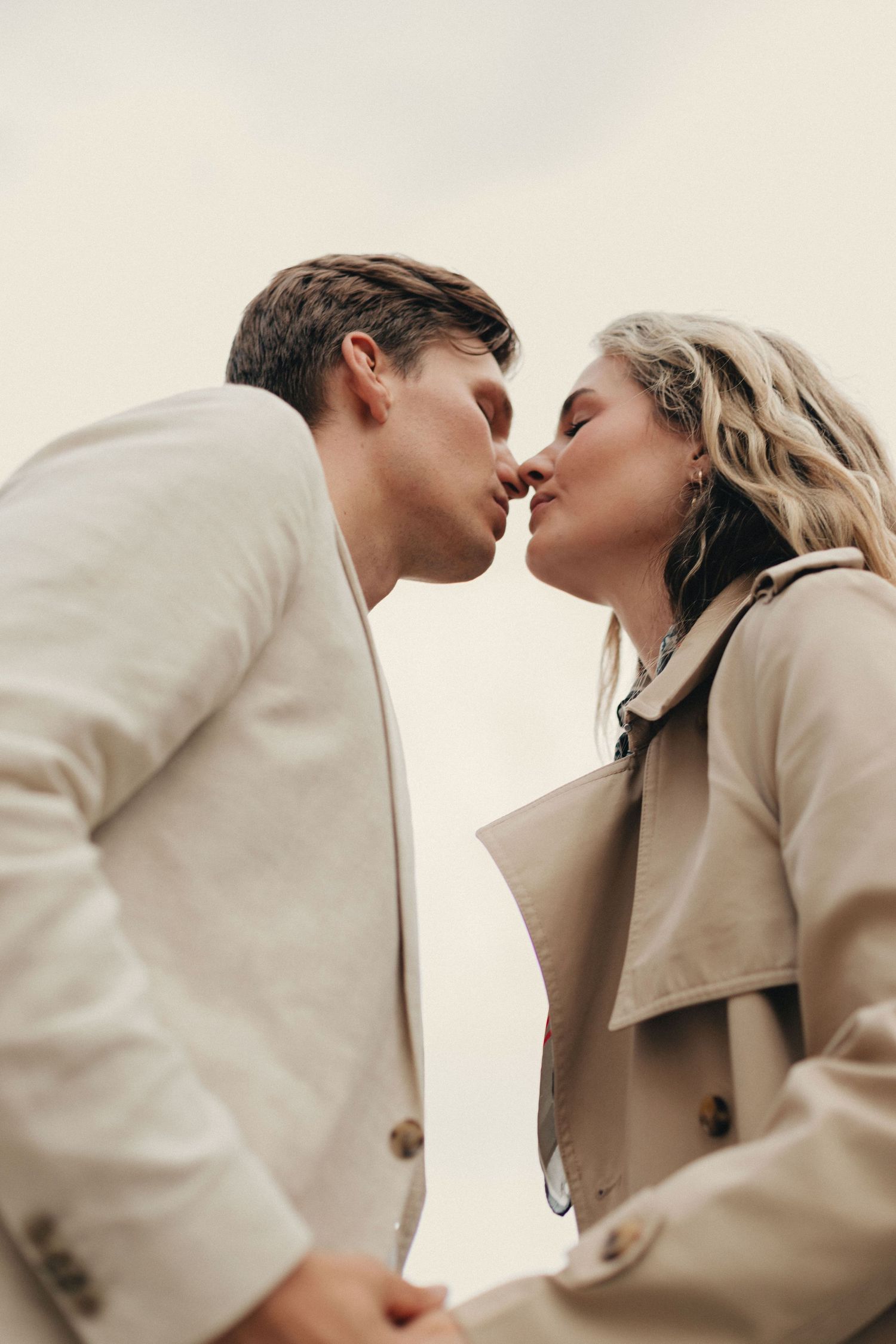 A romantic couple in beige coats share an intimate moment against a light background.