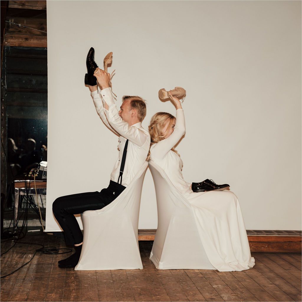 Couple in wedding attire playfully posing back-to-back while sitting and holding up beverages at a reception.