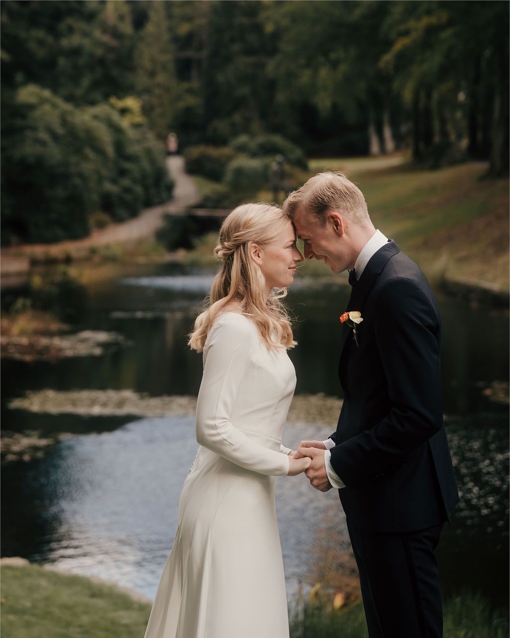 A couple in wedding attire share an intimate moment by a serene lakeside surrounded by trees during their outdoor ceremony.