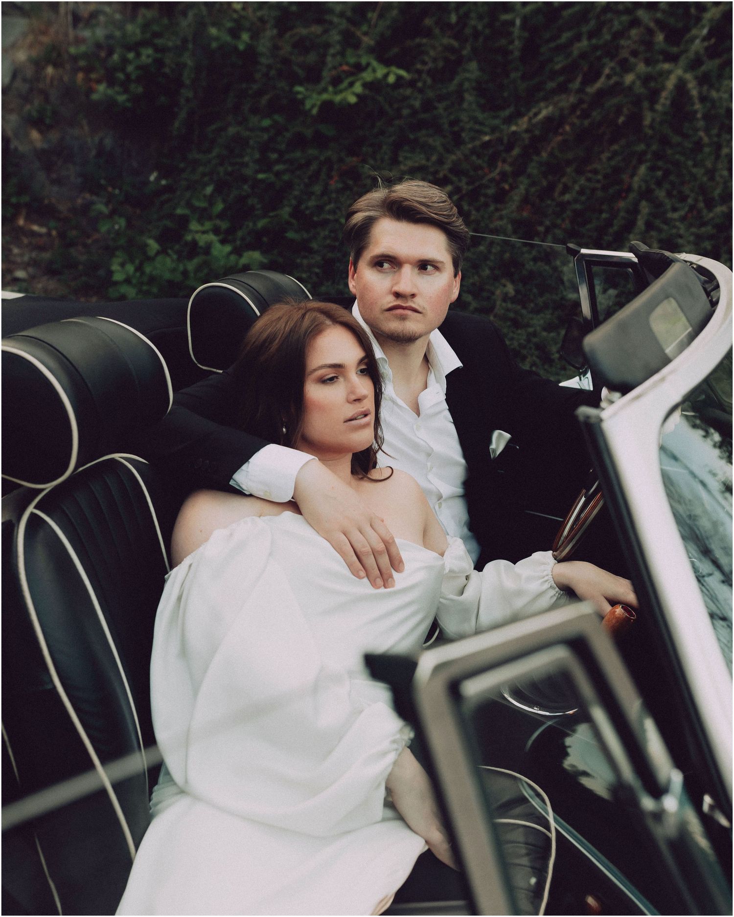 A couple in wedding attire share intimate moments in a vintage car during a moody outdoor photoshoot.