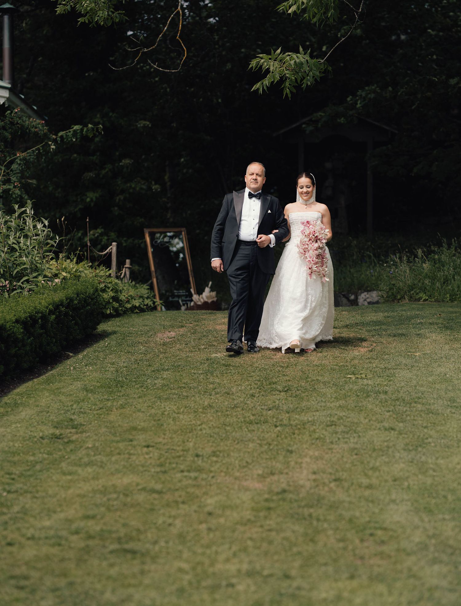 Bride and groom walking together on lush green lawn surrounded by trees during outdoor wedding ceremony.