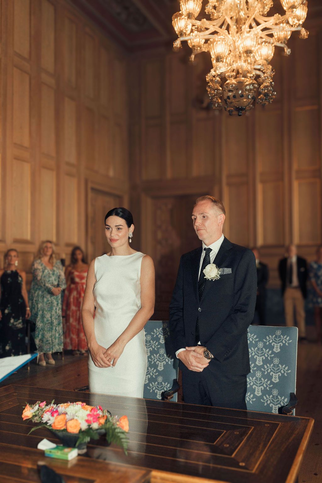 A couple stands together during a wedding ceremony in an elegant church with crystal chandeliers overhead.