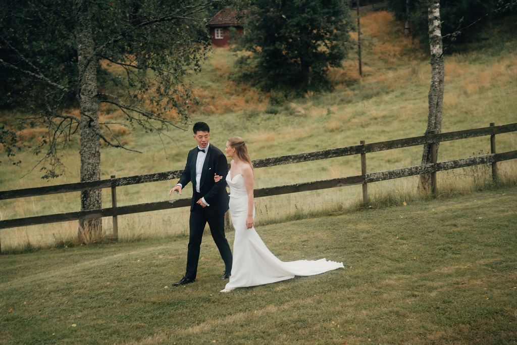 A newlywed couple poses together on a grassy hillside with a wooden ranch fence behind them.