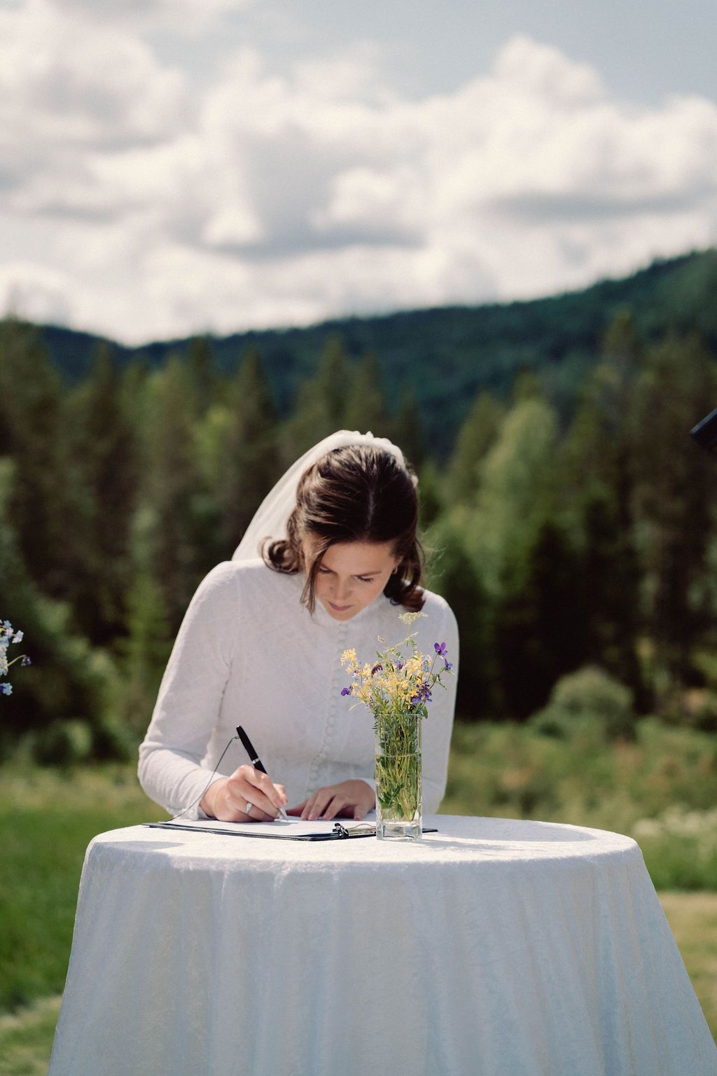Someone signs documents at a round table with white tablecloth and wildflower centerpiece outdoors in mountain setting.