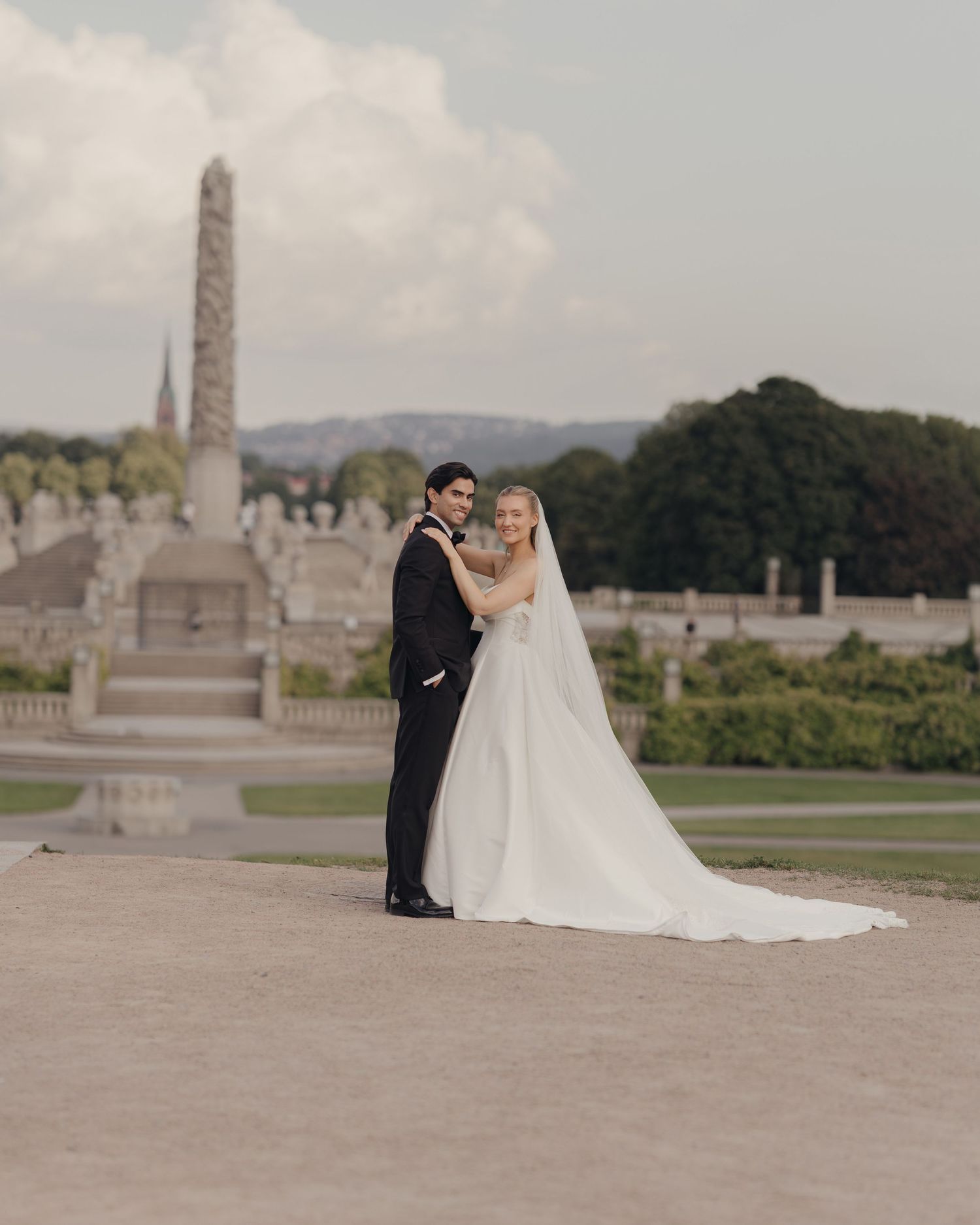 Bride in a flowing white gown and veil embraces her groom in a black tuxedo near a stone obelisk in a scenic park.