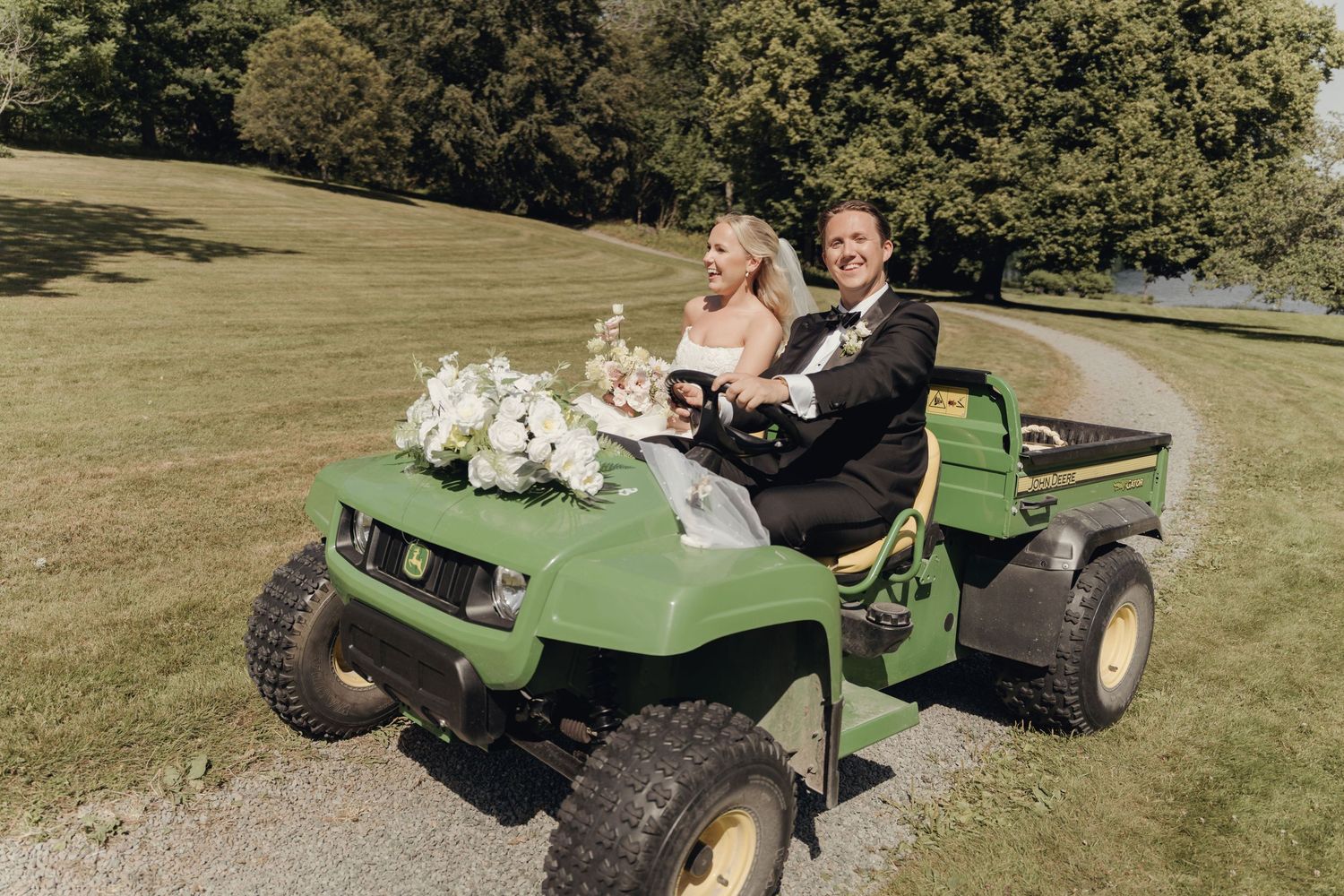 A bride and groom in wedding attire ride a green utility vehicle decorated with white flowers across a sunny grassy lawn.