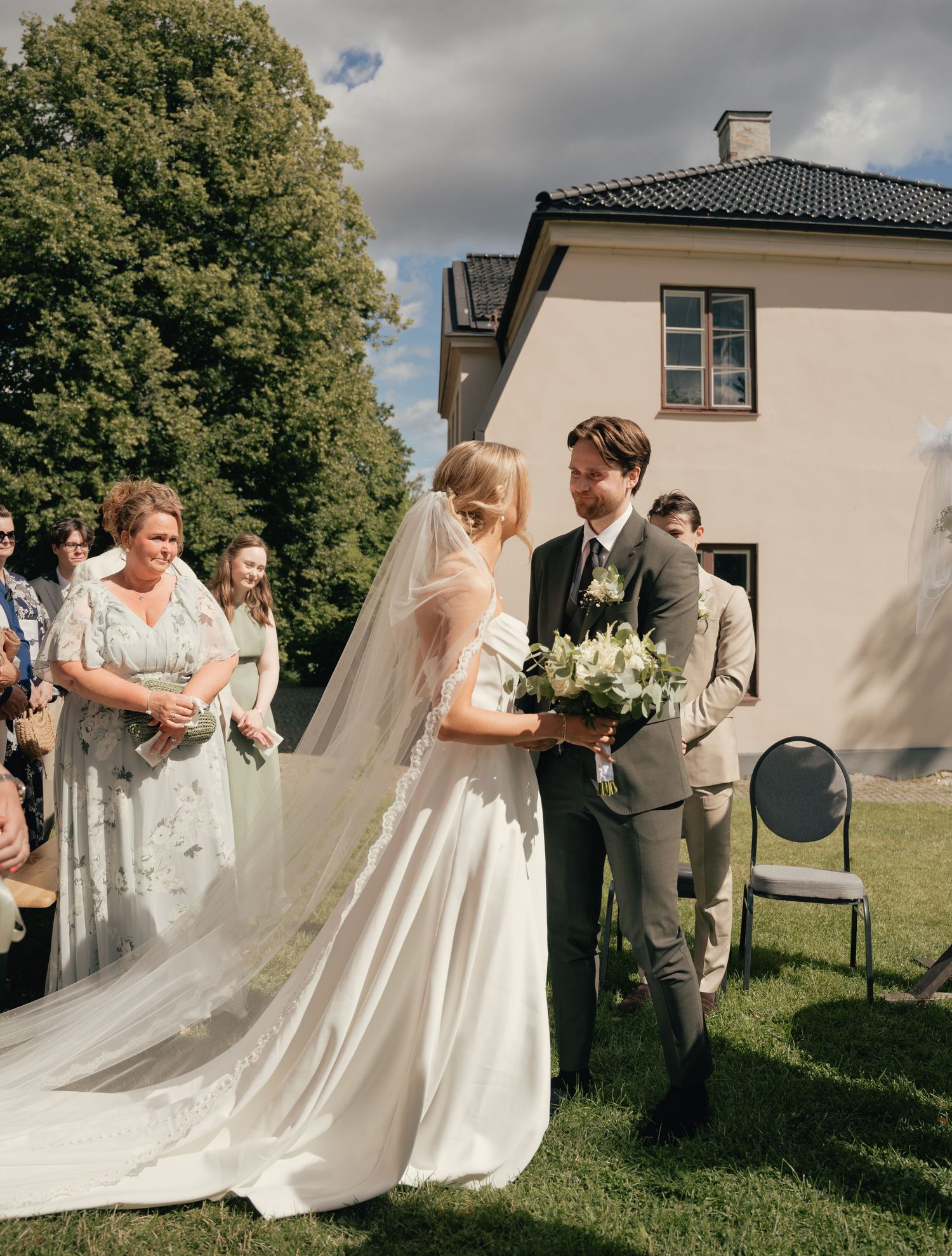 Bride in white gown with veil and bouquet walks with groom in gray suit at outdoor wedding ceremony.