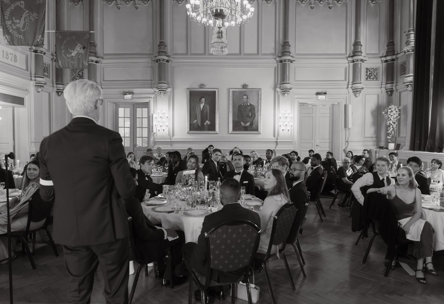 Black and white photo of a man standing to address guests seated at round tables in an elegant banquet hall with a chandelier.