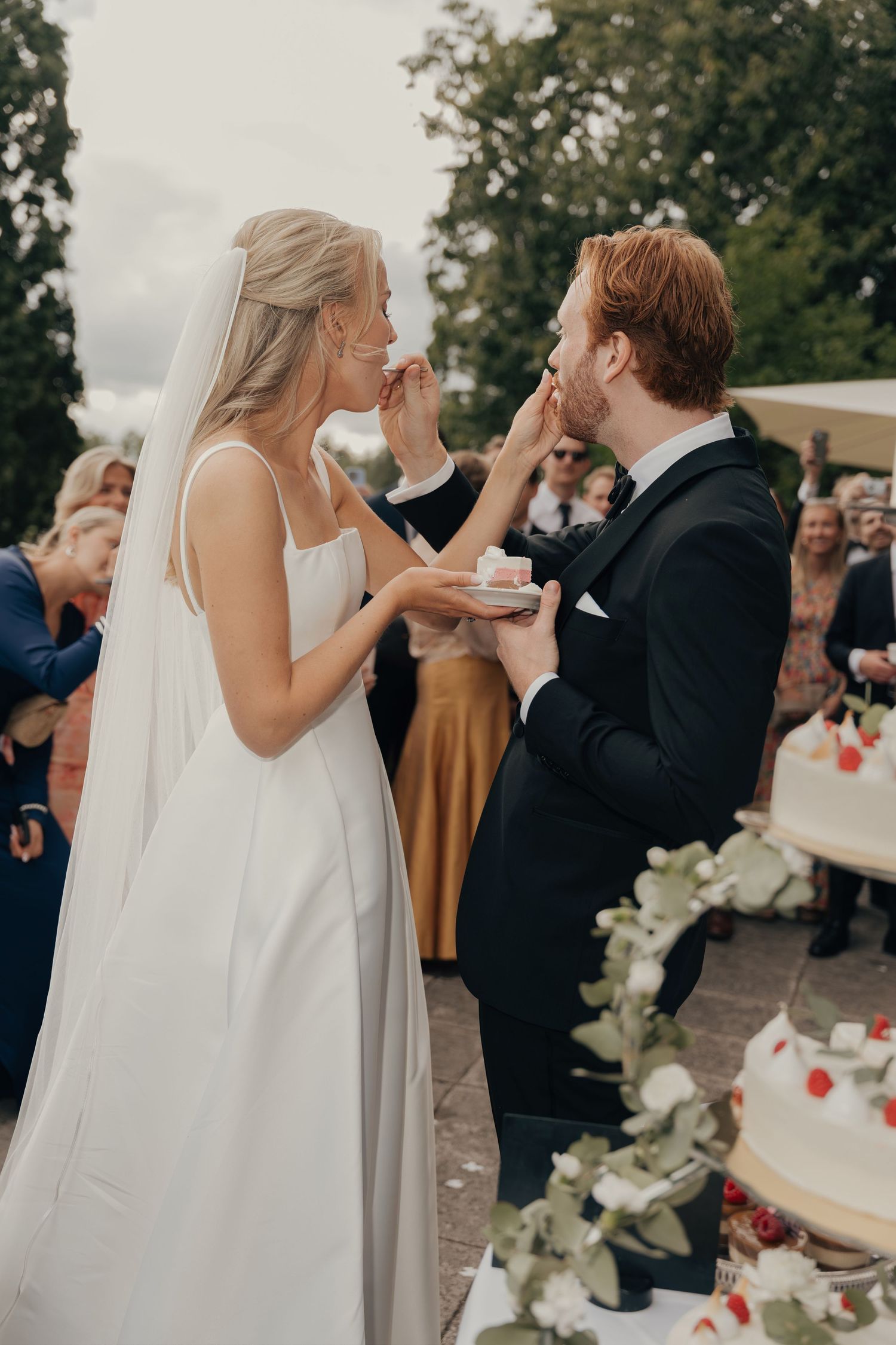 Bride and groom playfully feed each other wedding cake while guests watch at an outdoor reception.