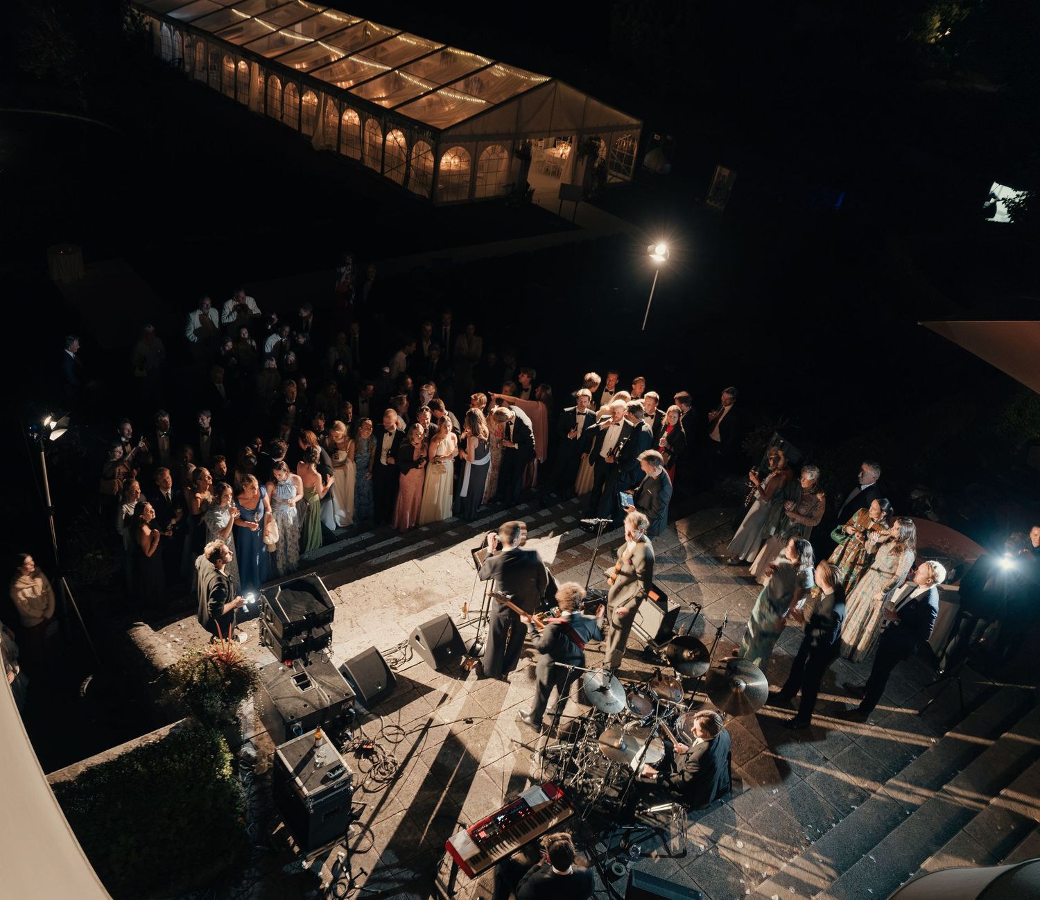 Wedding guests in formal attire gather around a live band performing on an outdoor terrace at night, with a glowing tent in the background.