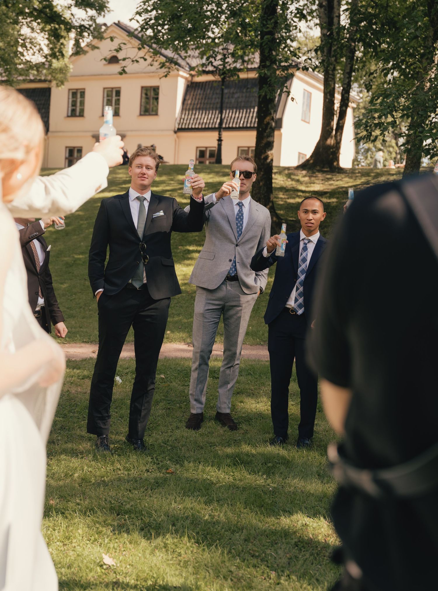 Groomsmen in suits raising bottles in a toast on a sunny outdoor wedding venue.